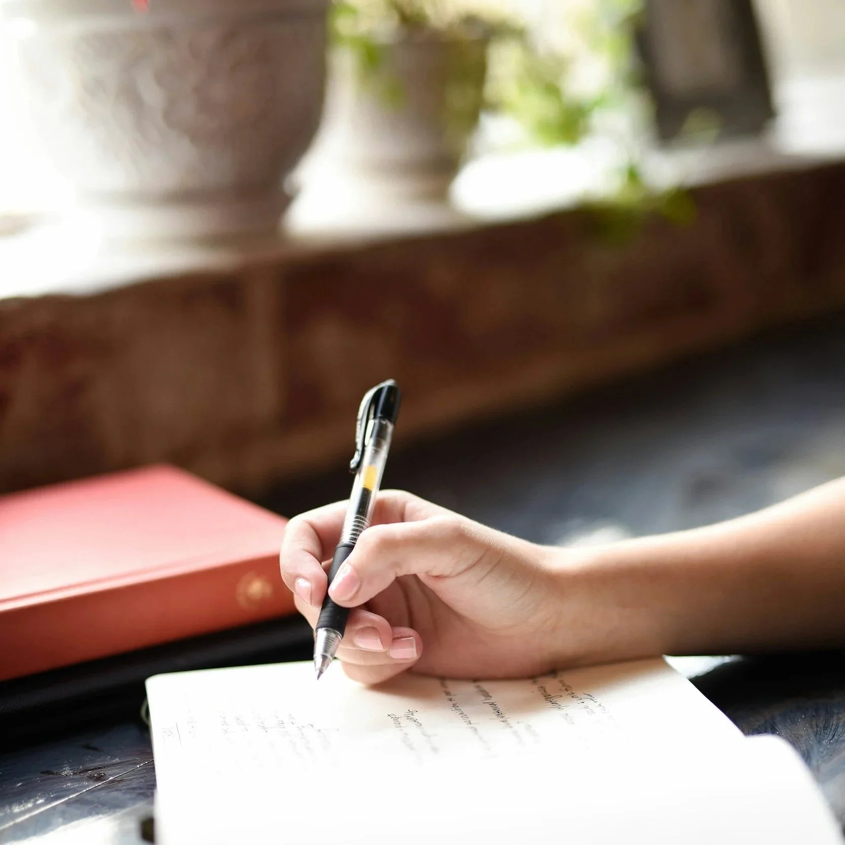 A person's hand holding a black pen writing on a notebook with handwritten notes, with a pink object and blurred potted plants in the background.
