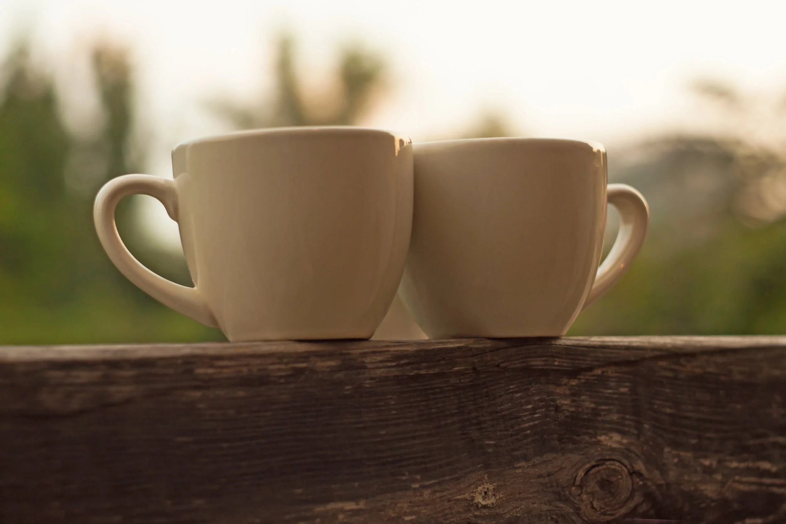 Two white ceramic coffee mugs on a wooden surface with a blurred outdoor background at sunrise or sunset.
