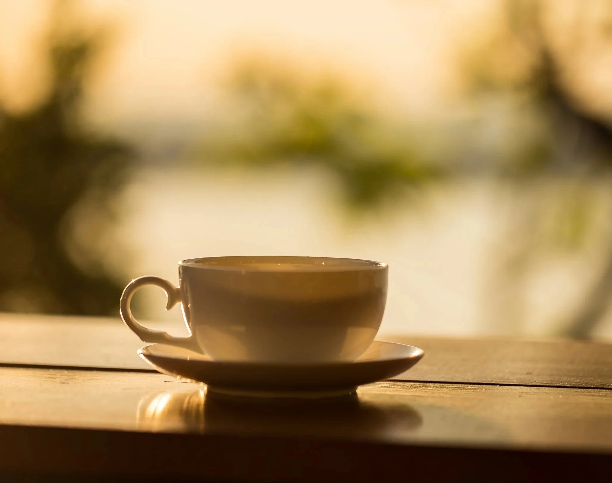 A white coffee cup with a handle sits on a matching saucer on a wooden surface. The background is blurred with warm tones and sunlight.