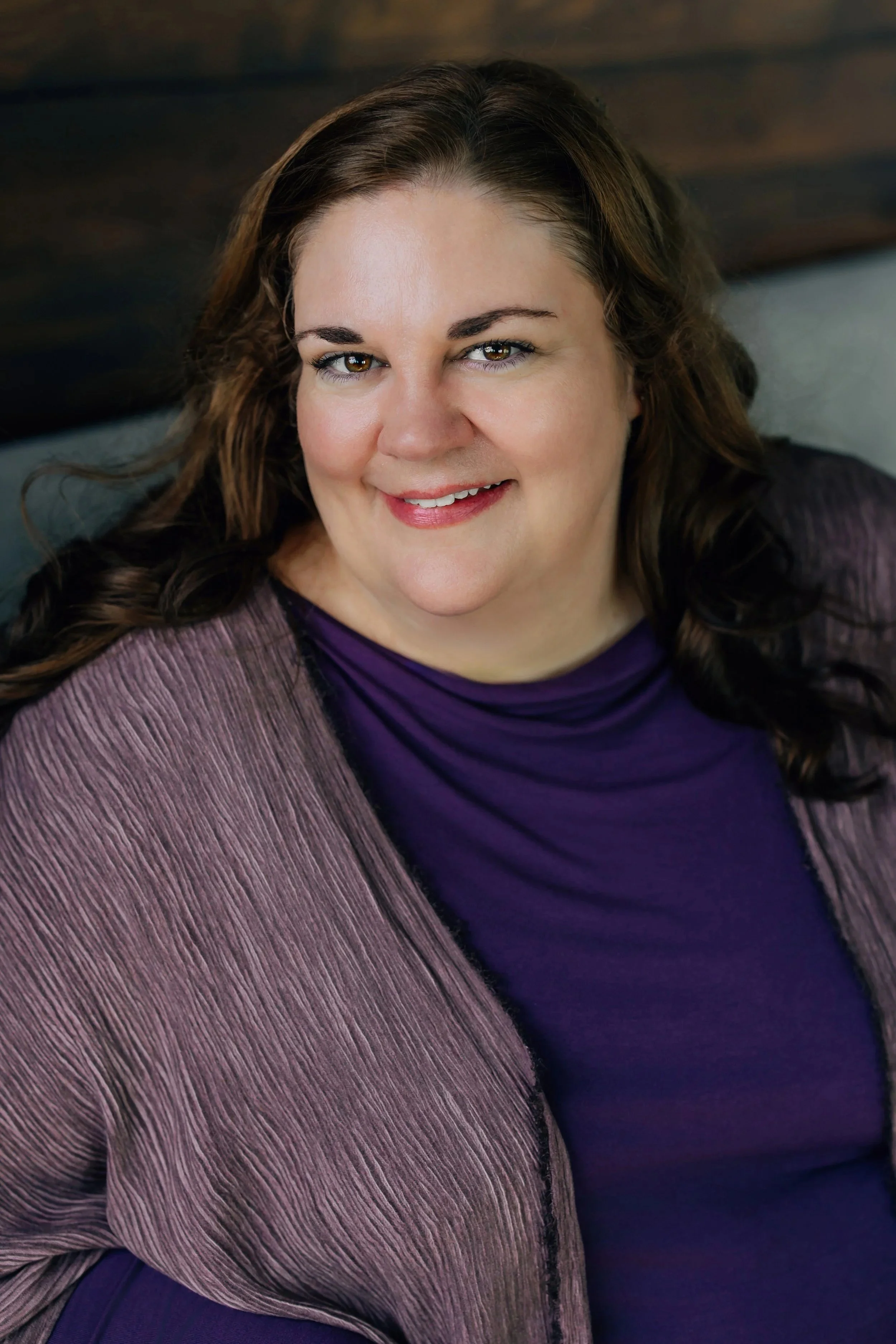 A woman with wavy brown hair smiling at the camera, wearing a purple shirt and a mauve jacket, sitting against a wooden background.