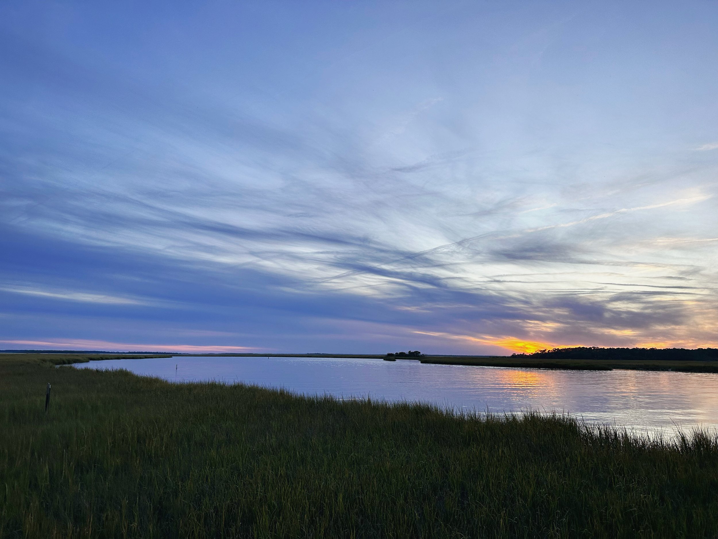 A landscape view of a river with grassy banks, a distant tree line, and a sky with blue and gray clouds at sunset.