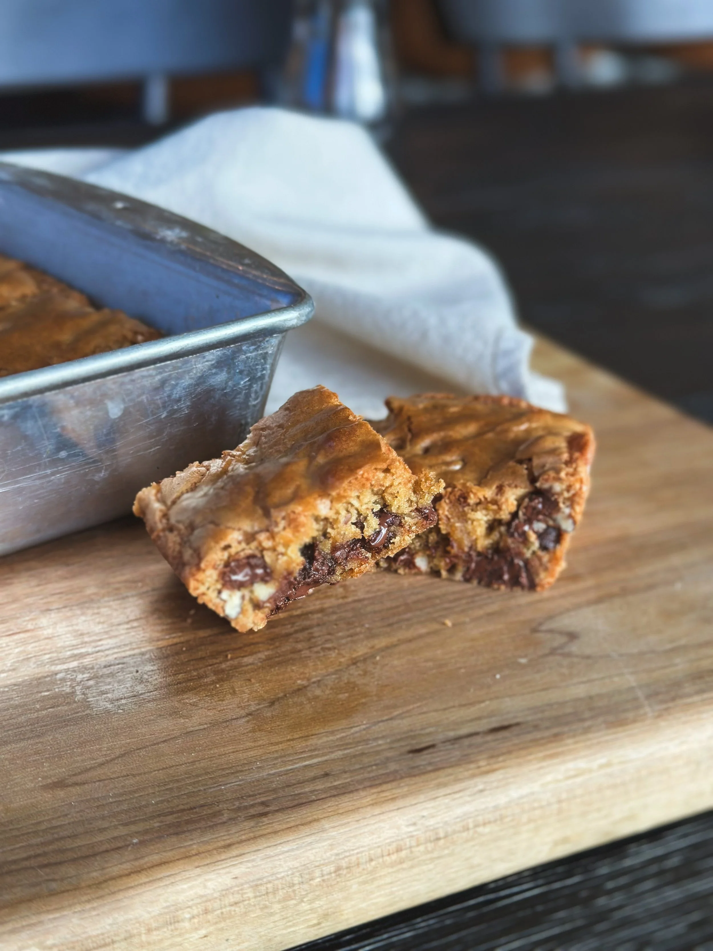 Close-up of a chocolate chip cookie bar with visible chunks of chocolate, placed on a wooden surface with a metal baking pan in the background.