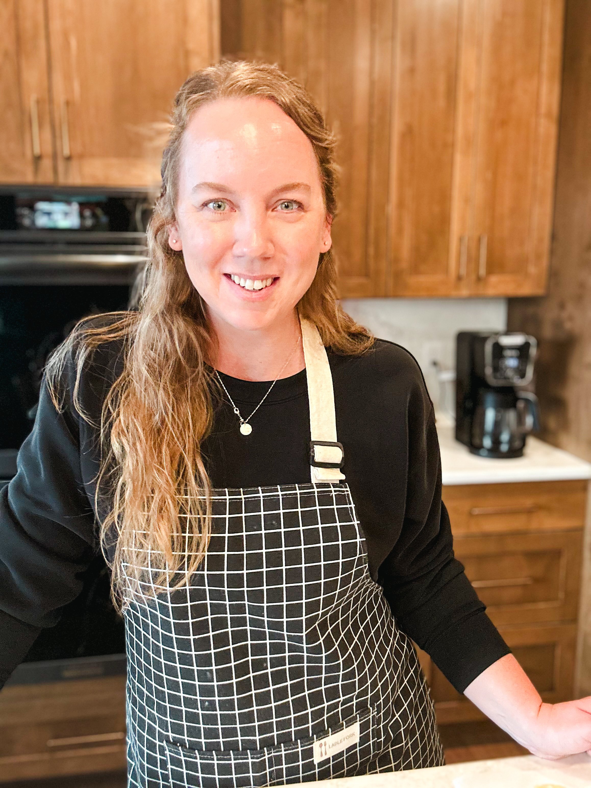 A woman wearing a black top, checkered apron, and necklace, standing in a kitchen with wooden cabinets and a coffee maker in the background.