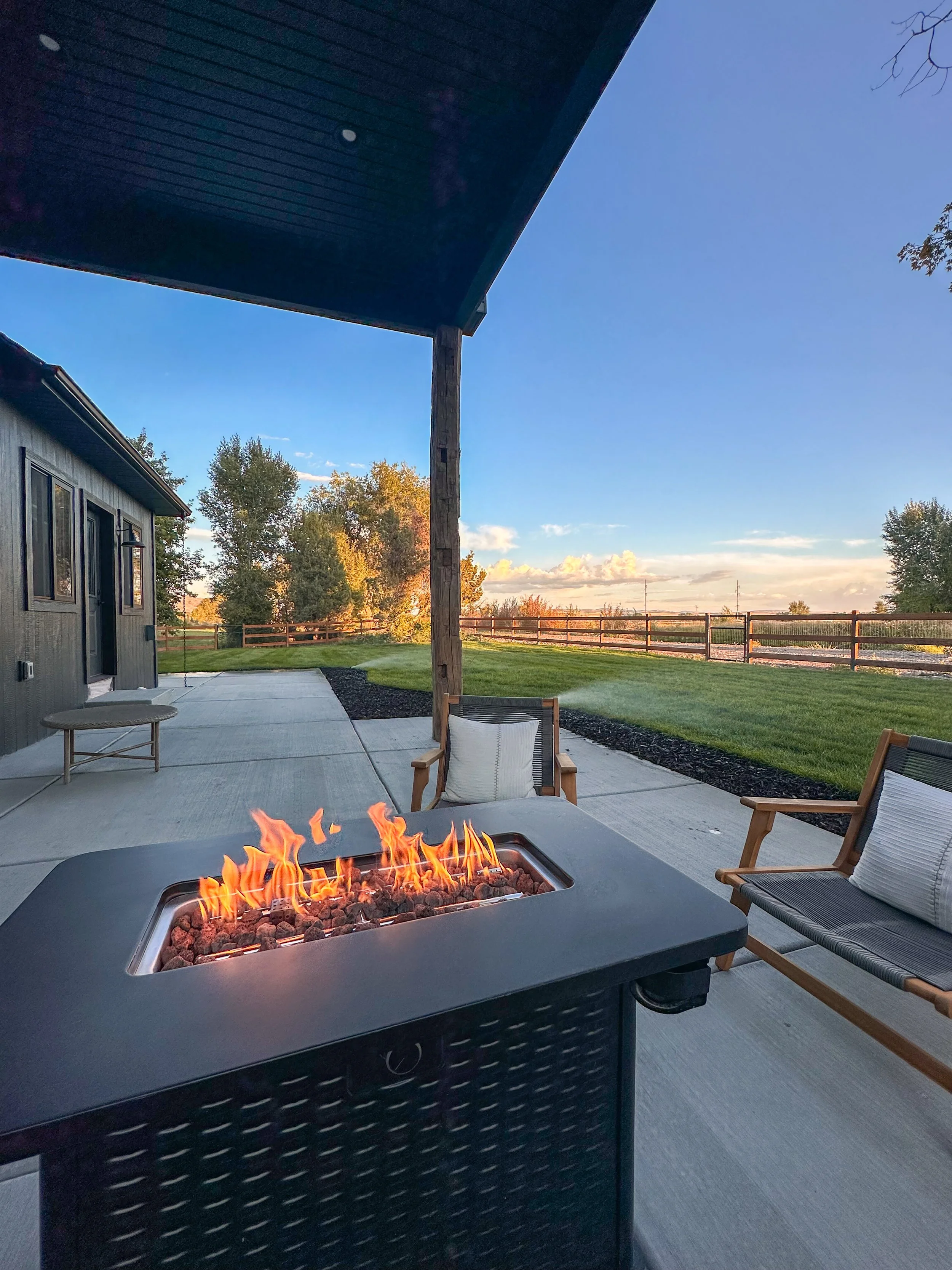 Outdoor patio with a fire table, two chairs with cushions, and a view of trees and a fence during sunset.