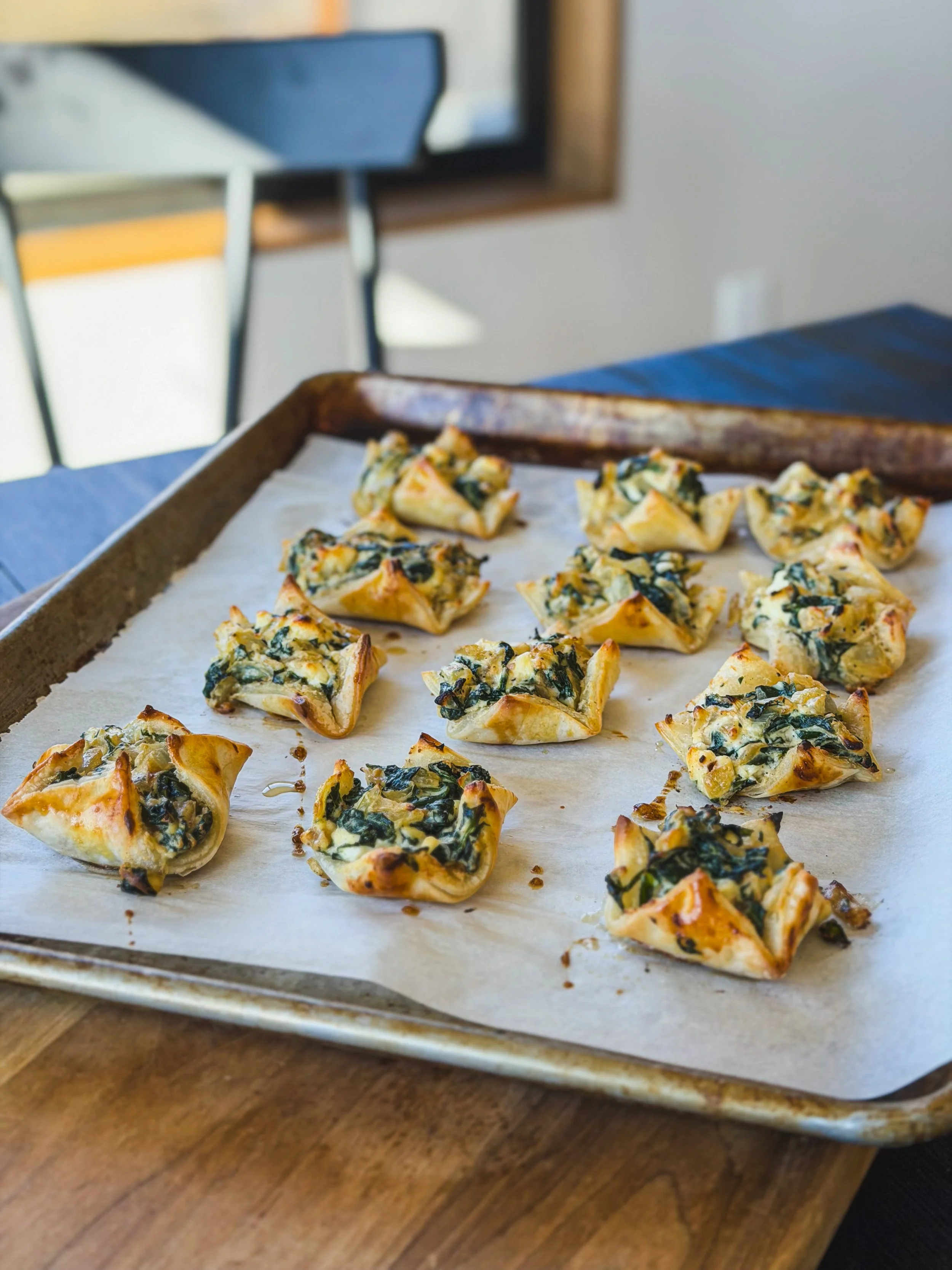 A baking sheet lined with parchment paper holding several spinach and cheese stuffed pastries or savory bites, with a blurred background of a kitchen or dining area.