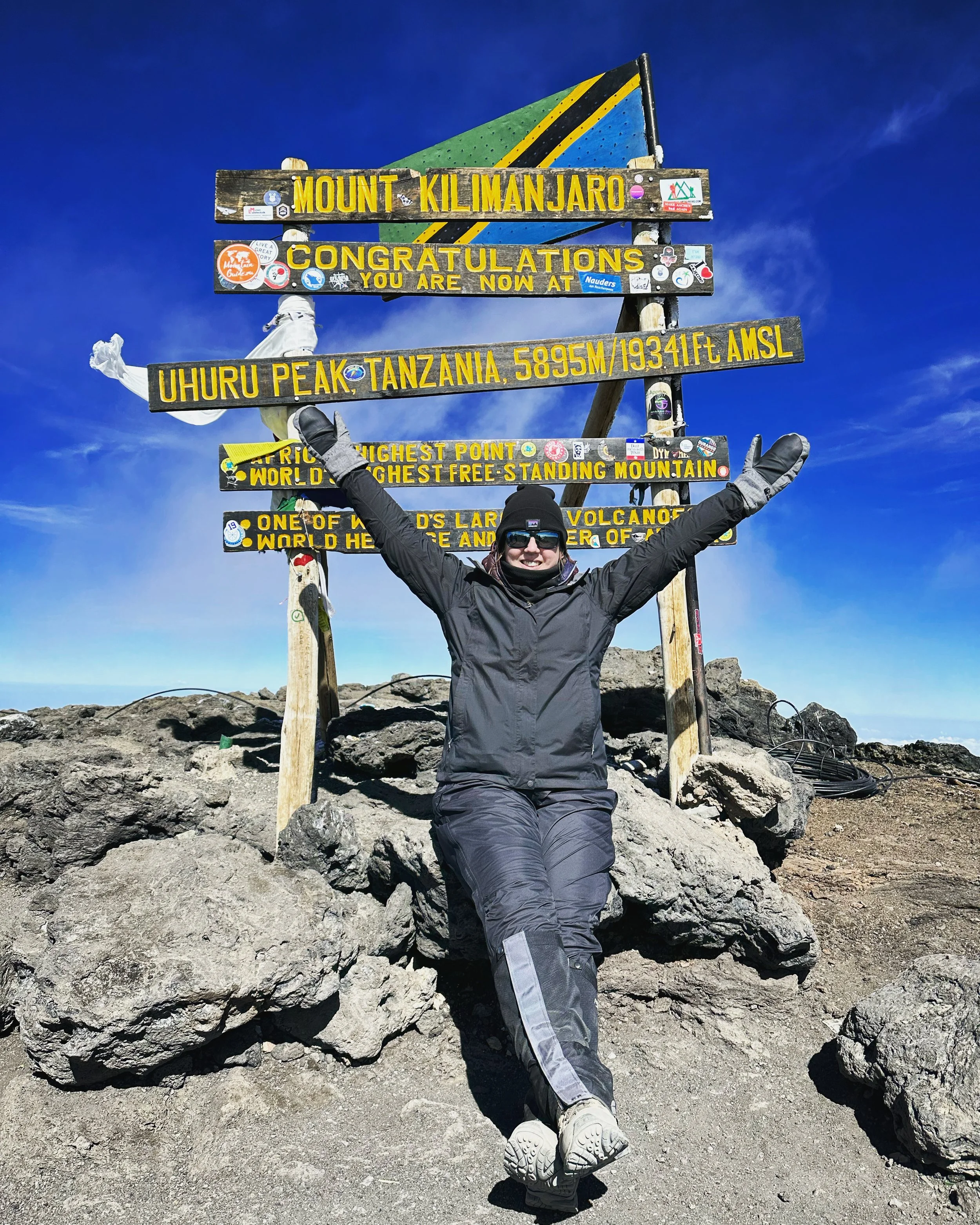 Person in black outdoor gear, wearing gloves and sunglasses, posing on rocky terrain at the summit of Mount Kilimanjaro in Tanzania. Signage shows the summit is at 5895 meters (19341 feet) above sea level, with a mountain flag behind it.