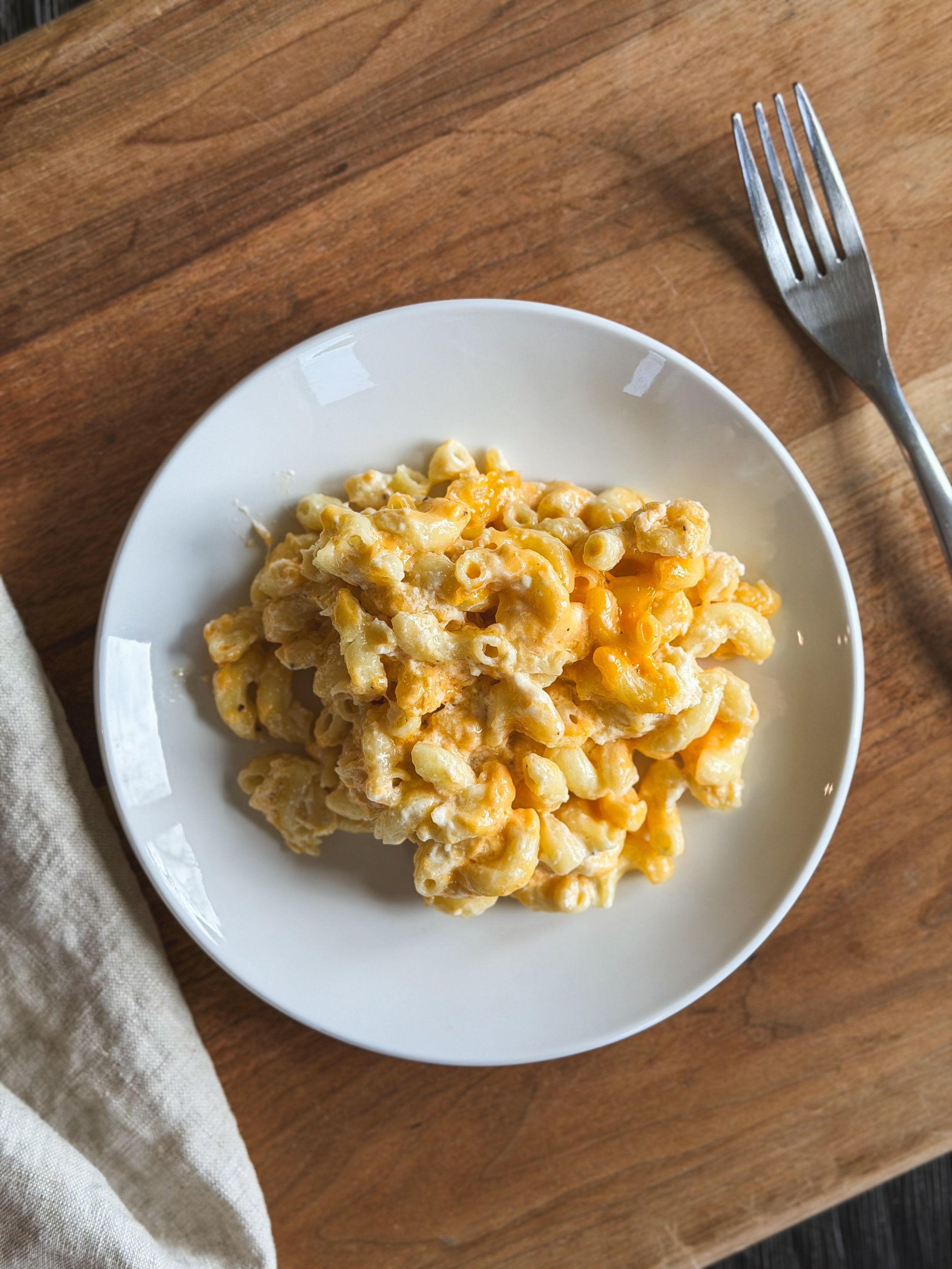 A plate of macaroni and cheese on a wooden table with a silver fork nearby and a beige cloth napkin in the corner.