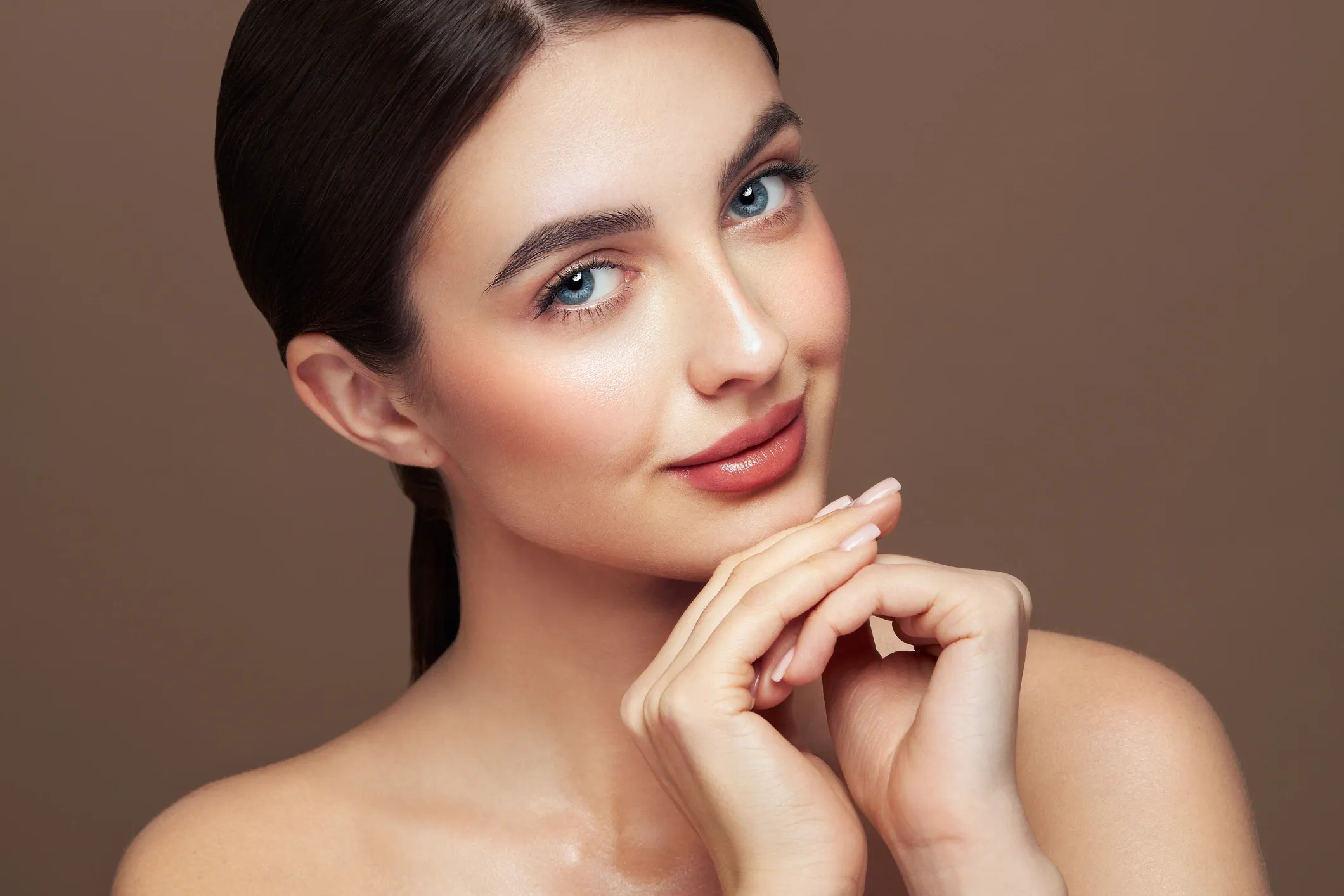 Close-up of a young woman with blue eyes and smooth skin, looking at the camera and smiling subtly, with her hand gently touching her chin against a plain brown background.
