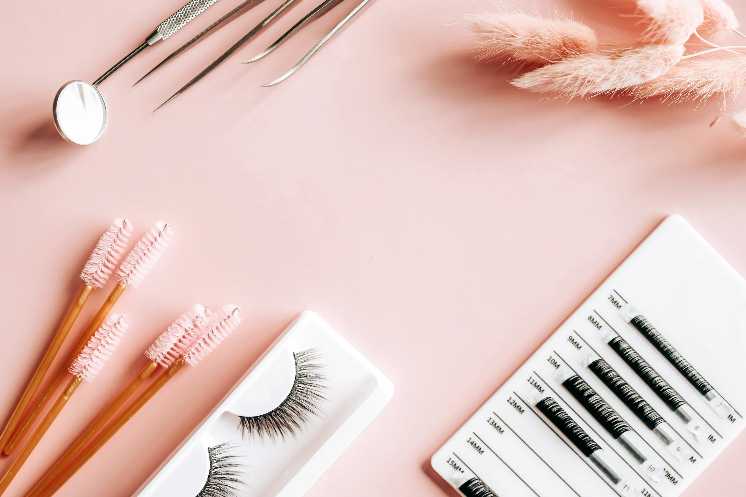Beauty tools including eyelash extension brushes, tweezers, a mirror, false eyelashes, and a lash extension sample card on a pink background with dried pink pampas grass.
