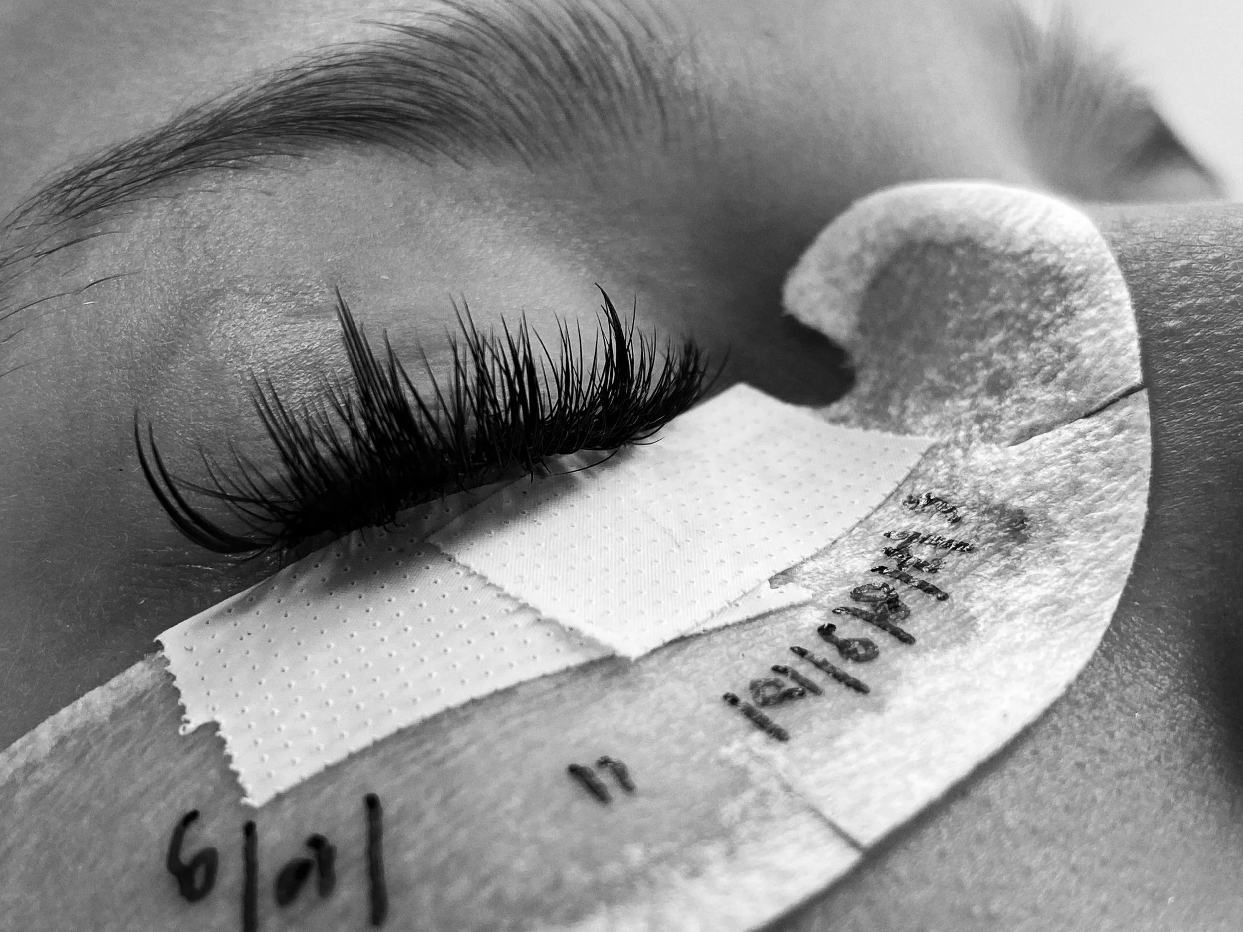 Close-up of closed eye with long, curled eyelashes and medical tape on eyelid, likely during eyelash extension procedure or eye treatment.
