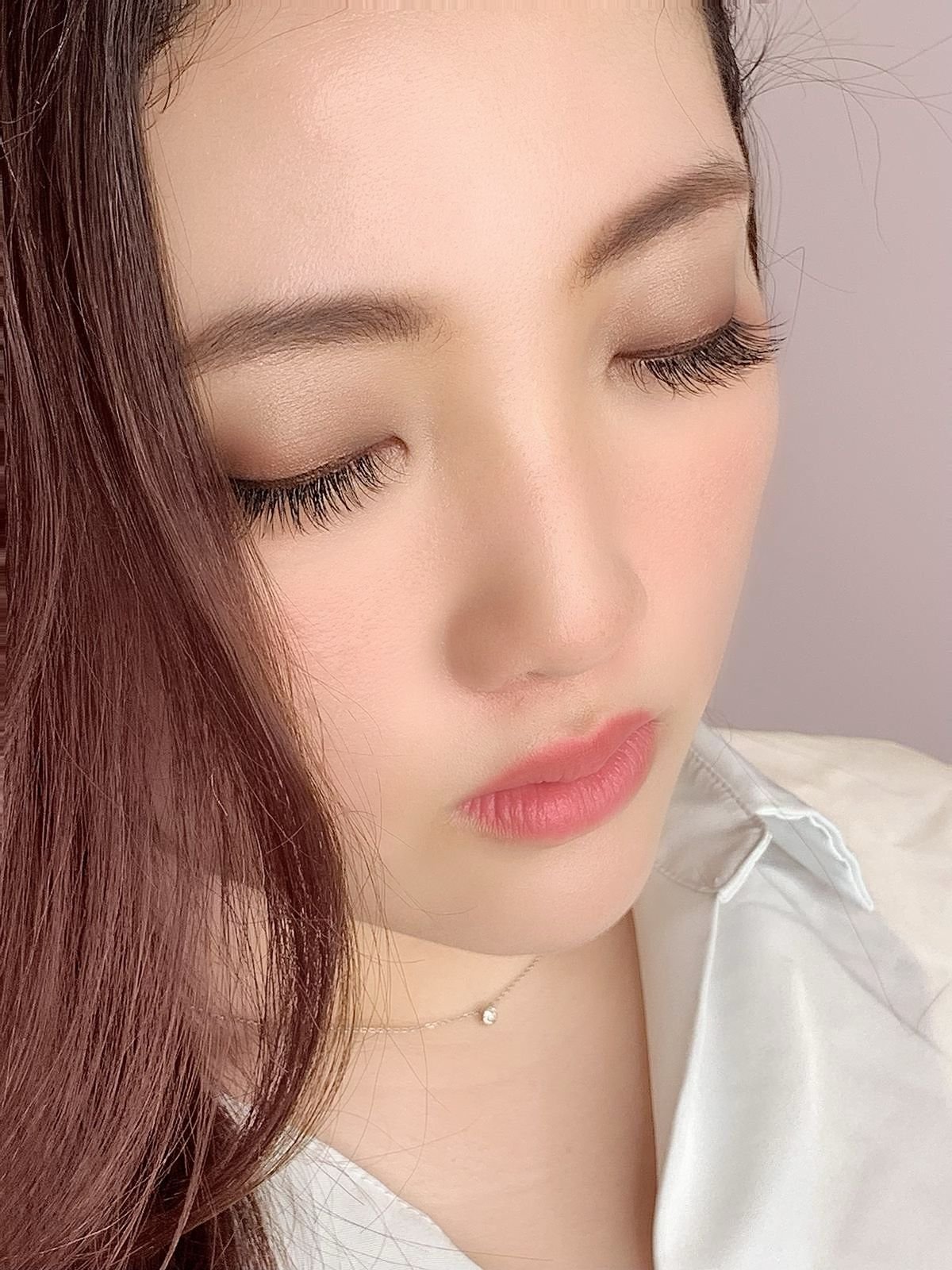 A close-up of a young woman with fair skin, closed eyes, long eyelashes, and light makeup, wearing a white collared shirt and a small necklace with a round pendant.