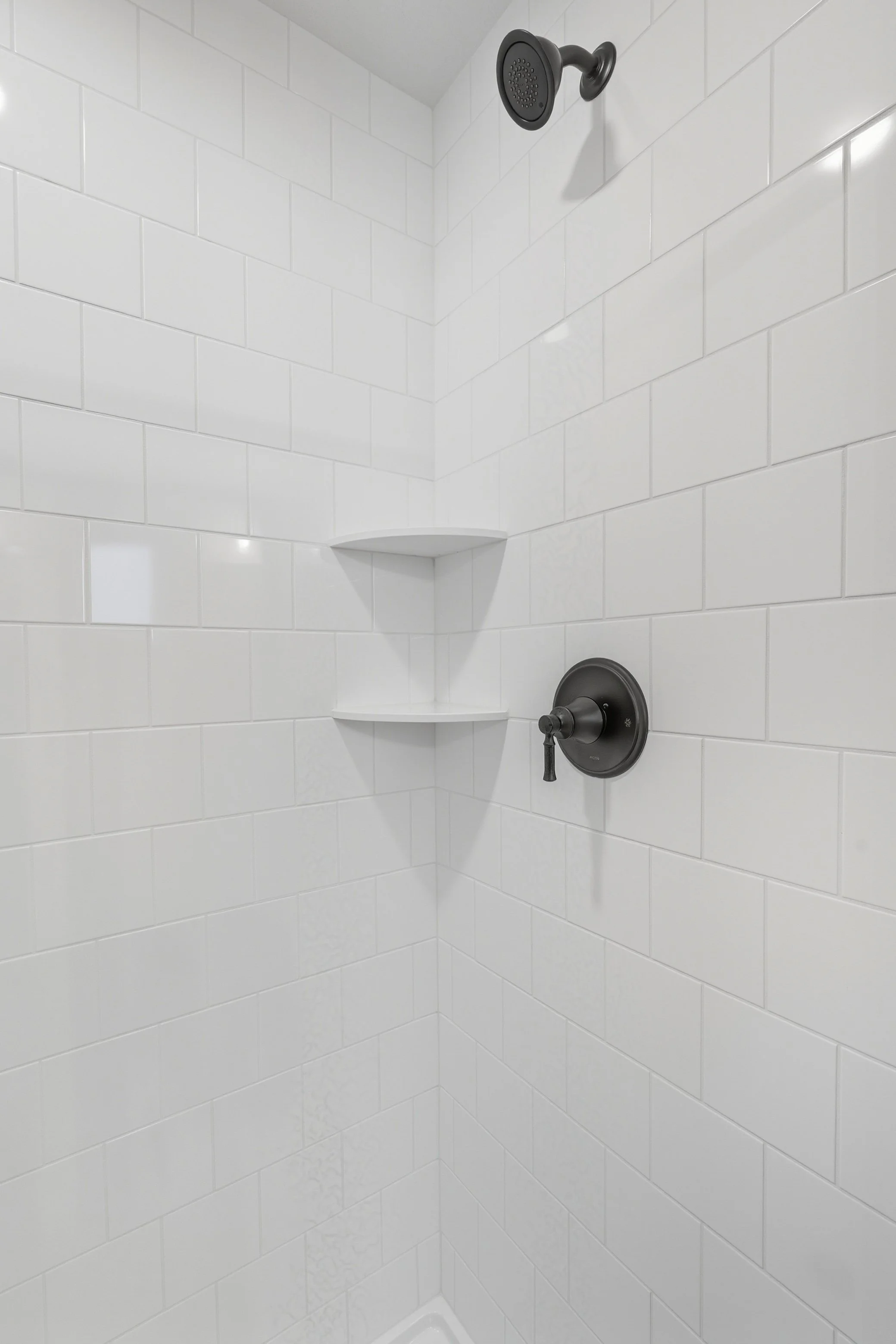 A white tiled shower with black fixtures, including a showerhead and a control valve, and two corner shelves.
