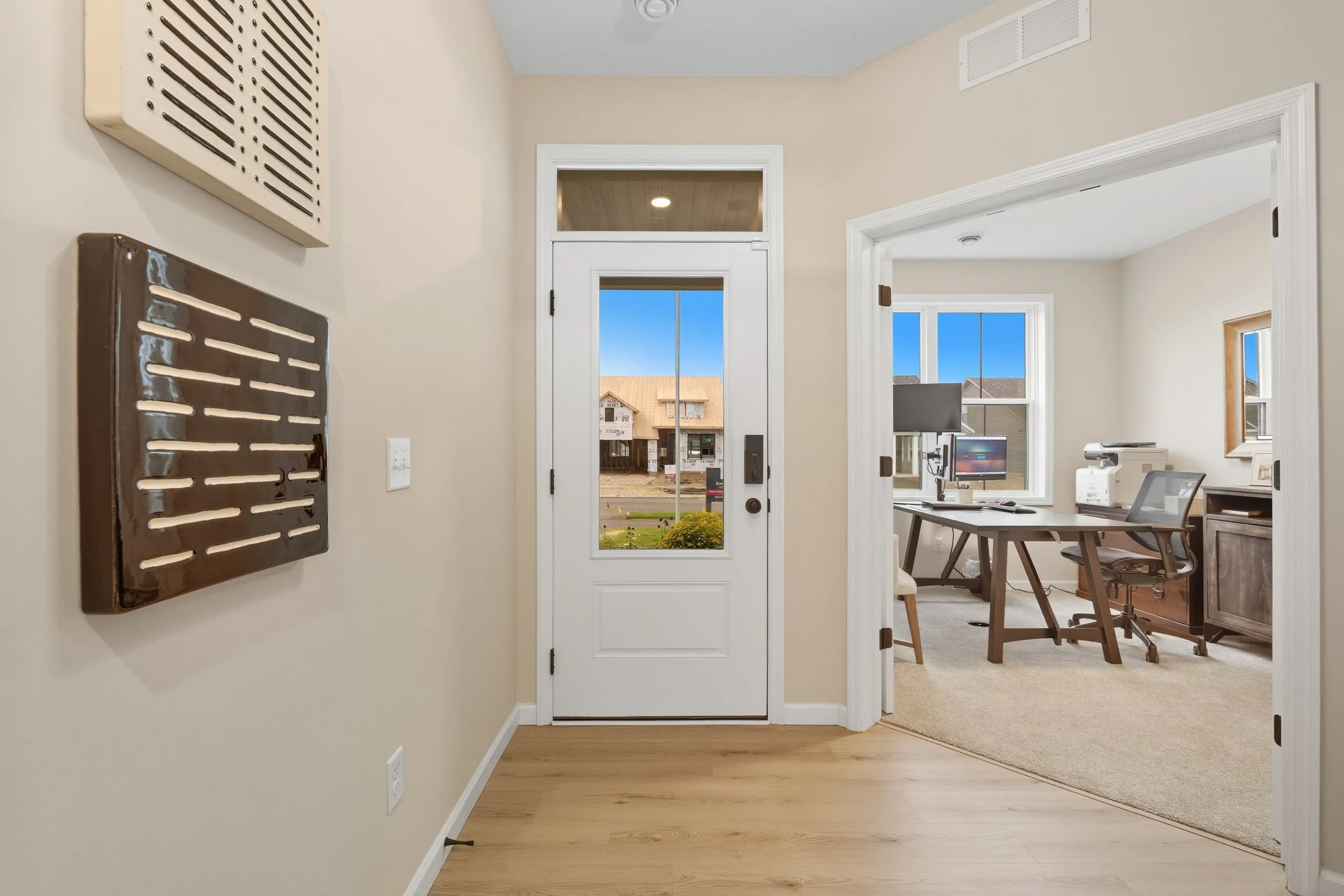 Entryway view of a home with a white front door and a view of houses outside. To the right, an office with a desk, chair, and computer setup is visible through a doorway.