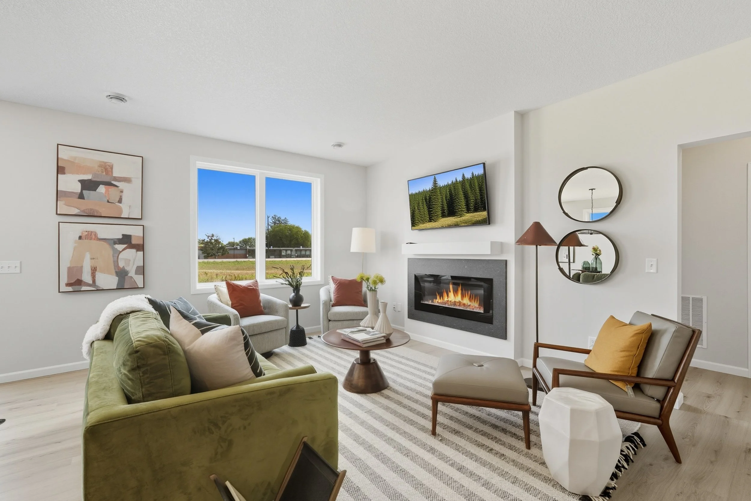 Living room with modern decor featuring a green sofa, a fireplace, a wall-mounted TV, abstract wall art, and large windows showing an outdoor view.