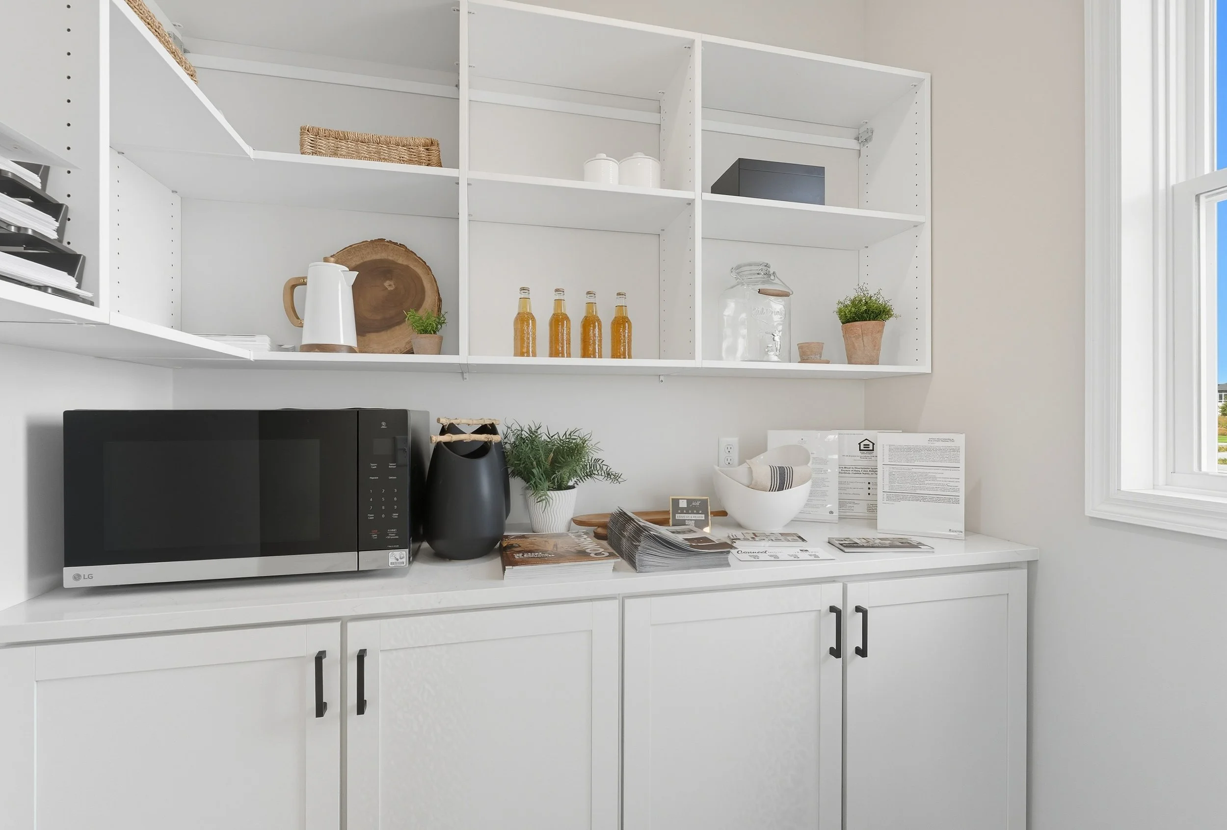 Kitchen countertop with microwave, potted plants, jars, and magazines, with open shelving above holding decorative items and storage boxes.