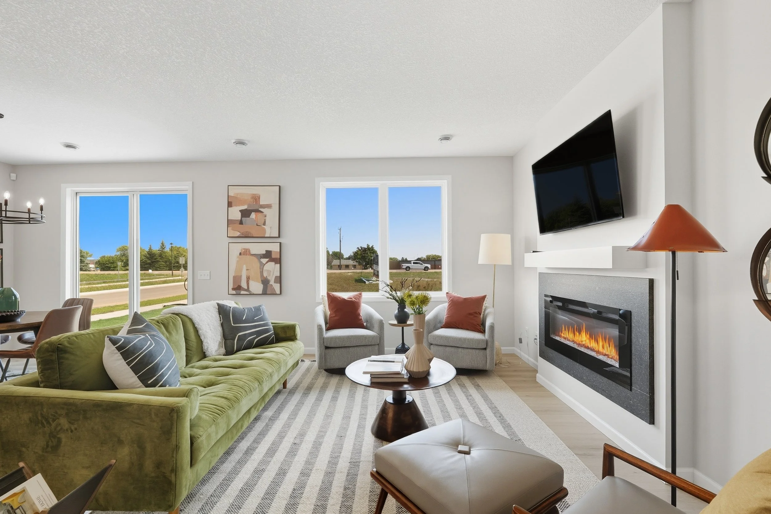 Living room with green sofa, two gray armchairs, fireplace, wall-mounted TV, and large windows showing blue sky and a street scene.
