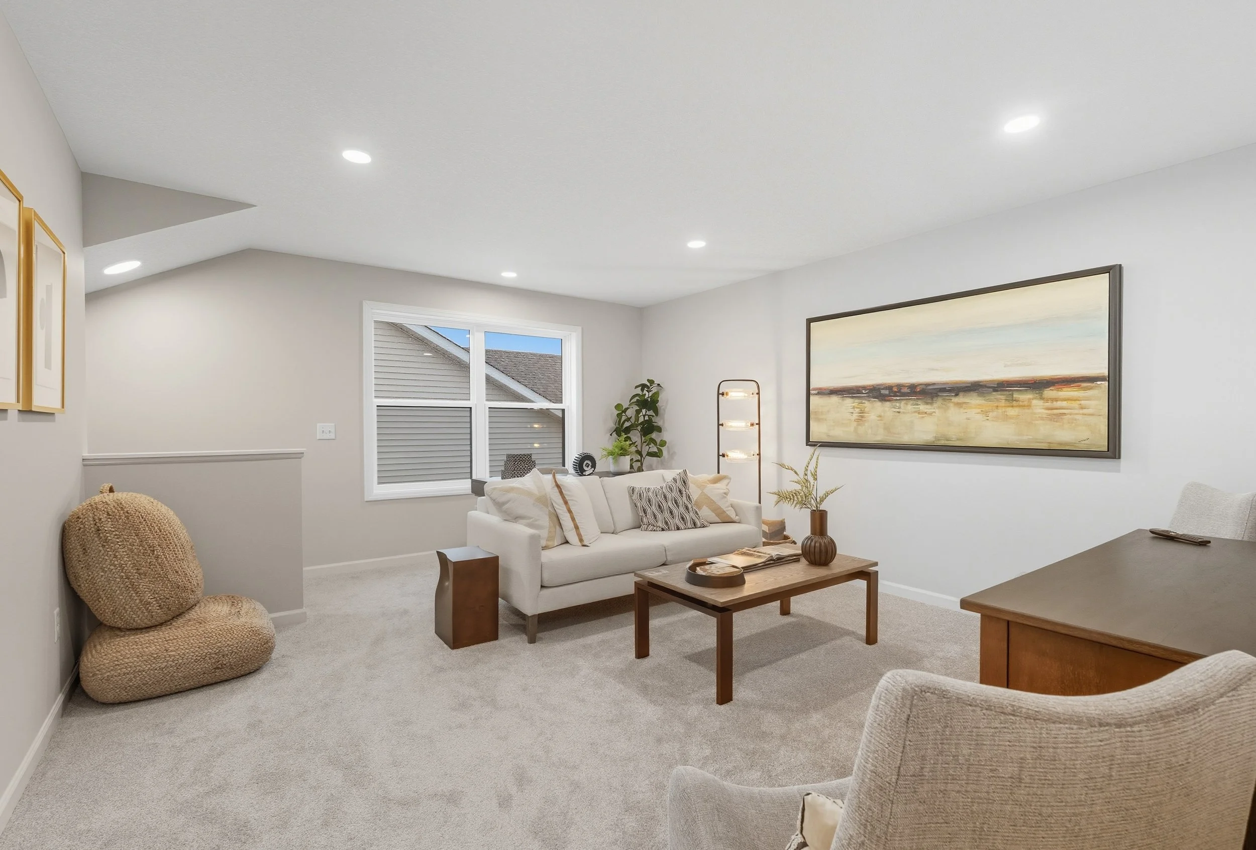 A living room with a white sofa, beige armchair, wooden coffee table, large landscape painting, window, and various decor items including a floor lamp and potted plants.
