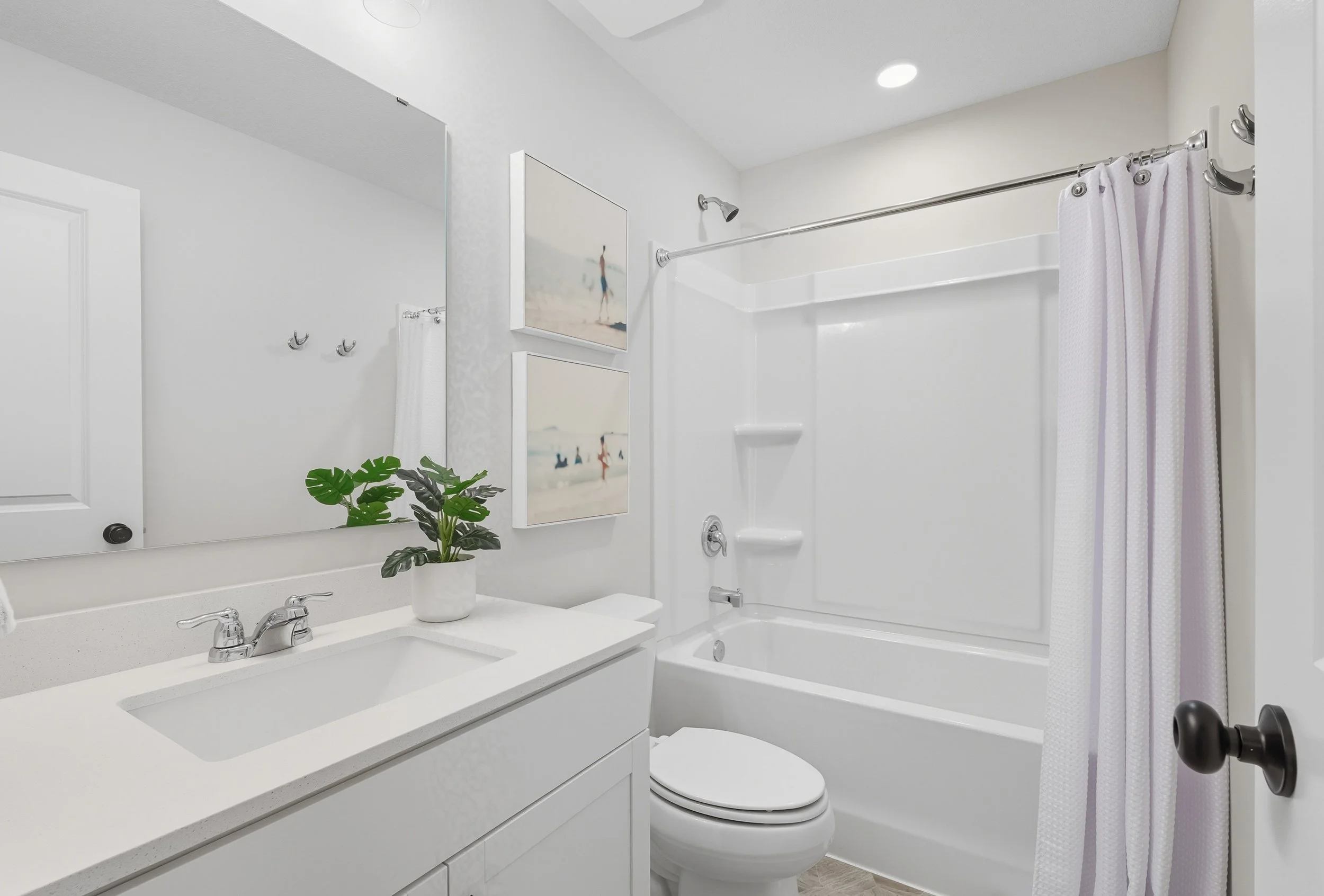 A clean, white bathroom with a vanity mirror, a potted plant, a sink, a toilet, a bathtub with a shower, and framed beach-themed artwork on the wall.