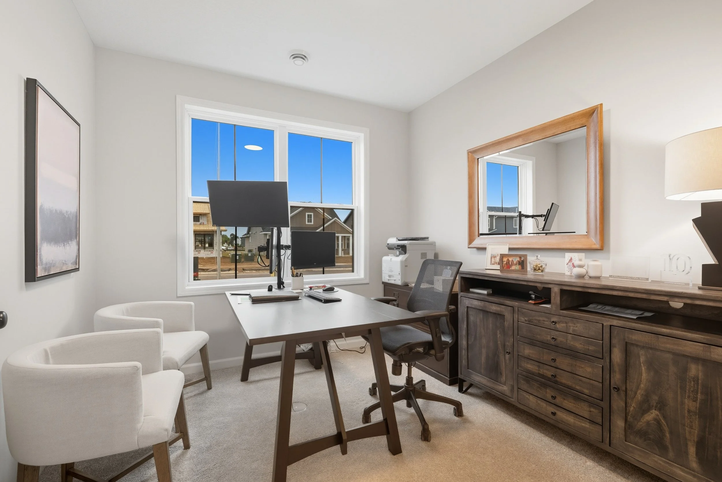 Home office with a desk, multiple monitors, chairs, and a large mirror on the wall, with a window showing a neighborhood outside.