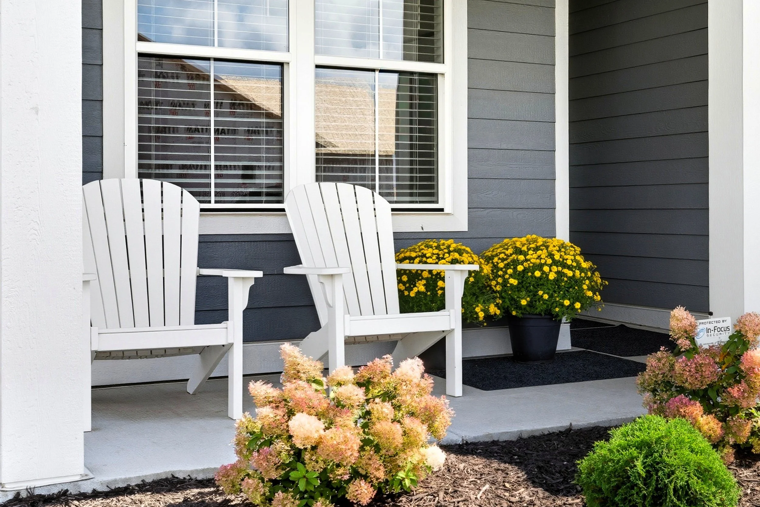 A front porch with two white Adirondack chairs, yellow potted flowers, pink hydrangeas, a small green shrub, and a window with white trim on a house with gray siding.