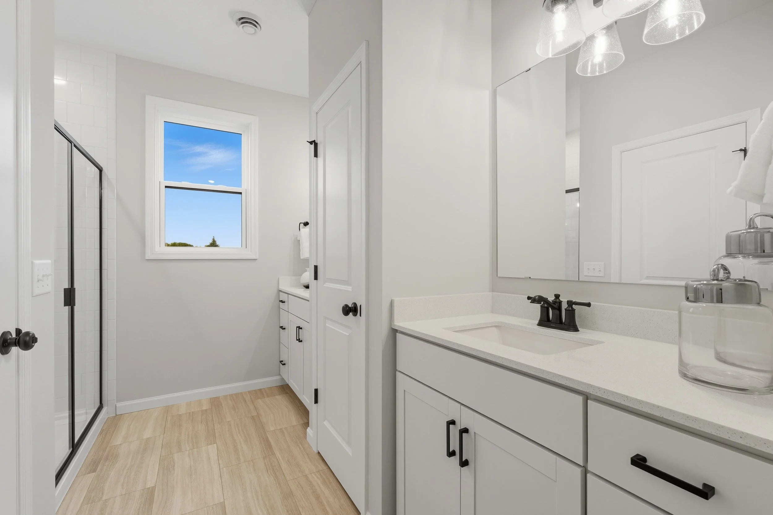 A modern bathroom with white cabinets, a large mirror, black fixtures, a window showing a blue sky, and a glass shower door.