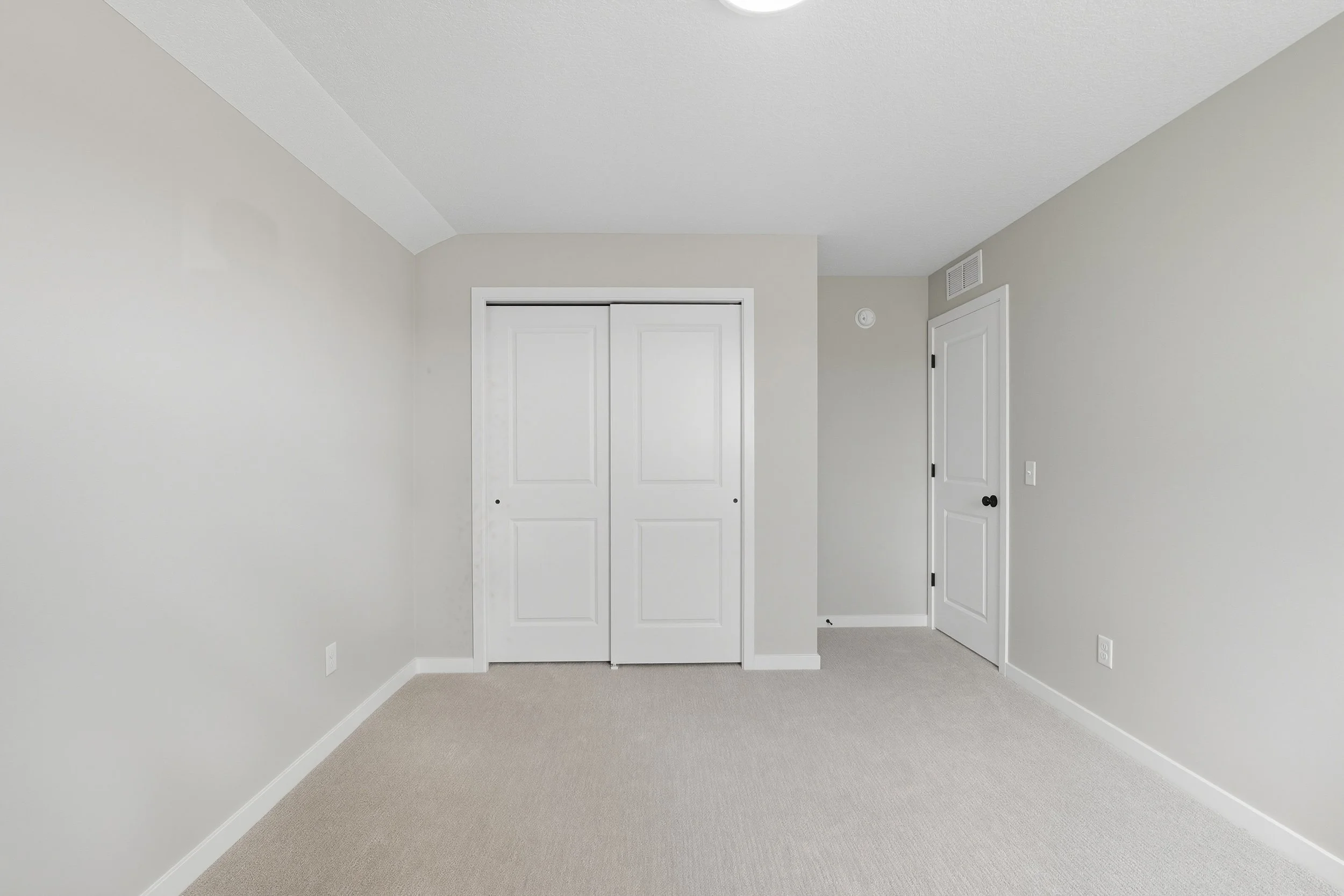Empty room with beige carpet, light gray walls, white trim, a closet with white double doors, a door with a black doorknob, and a ceiling vent.