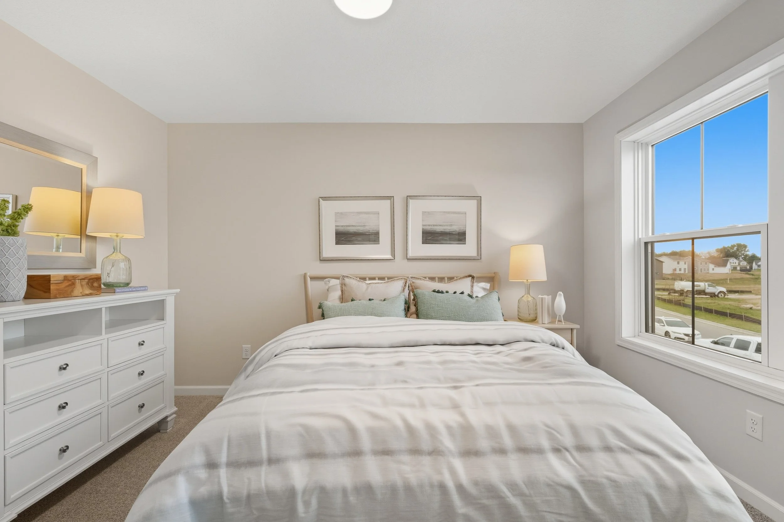 A cozy bedroom with a large bed dressed in white linens and pillows, flanked by two nightstands with lamps, a dresser on the left, and a large window showing a neighborhood outside.