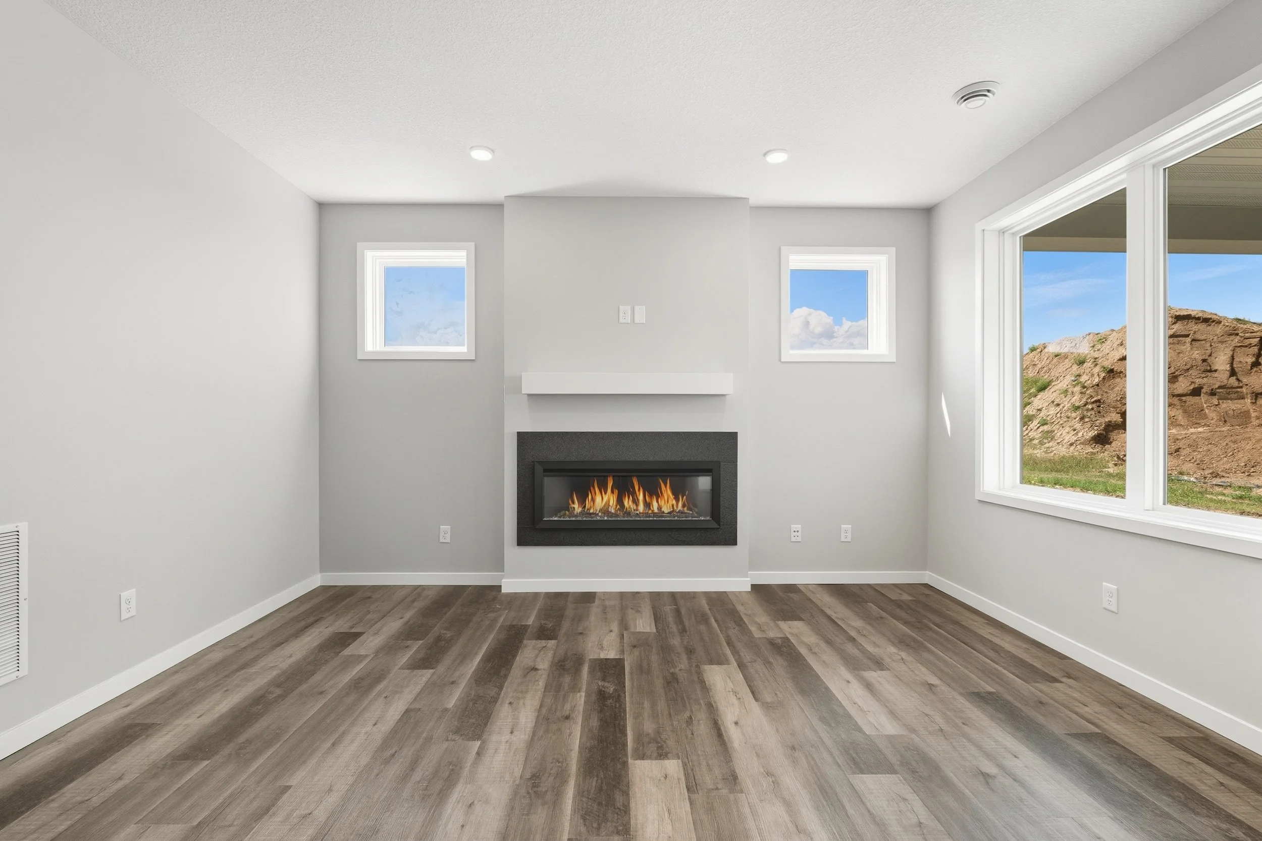 Empty living room with white walls, wood flooring, a modern fireplace, and large windows showing a landscape outside.
