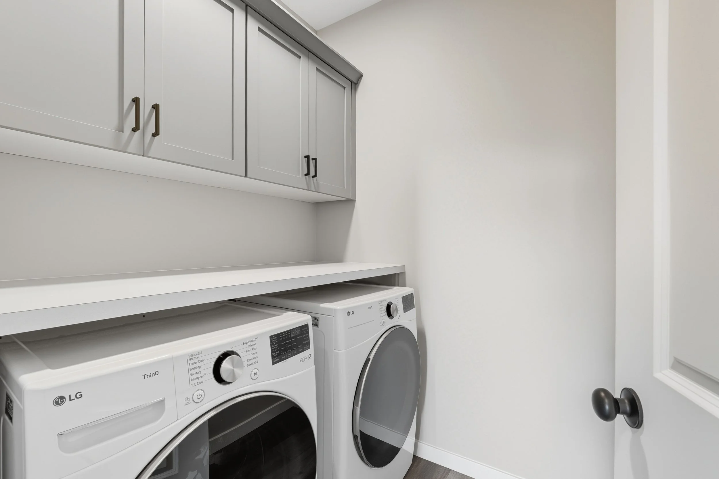 A laundry room with a white washing machine and dryer placed side by side, above which are grey cabinets, and a door with a black round handle on the right side.