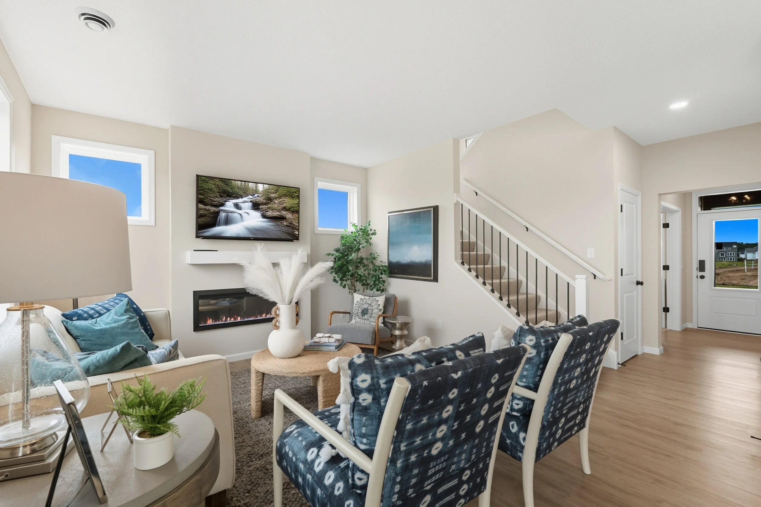 Living room with beige walls, hardwood floor, blue patterned chairs, a beige sofa, a round wicker coffee table, a wall-mounted flat-screen TV showing a waterfall scene, a modern fireplace, a potted plant, and a staircase with black railings.