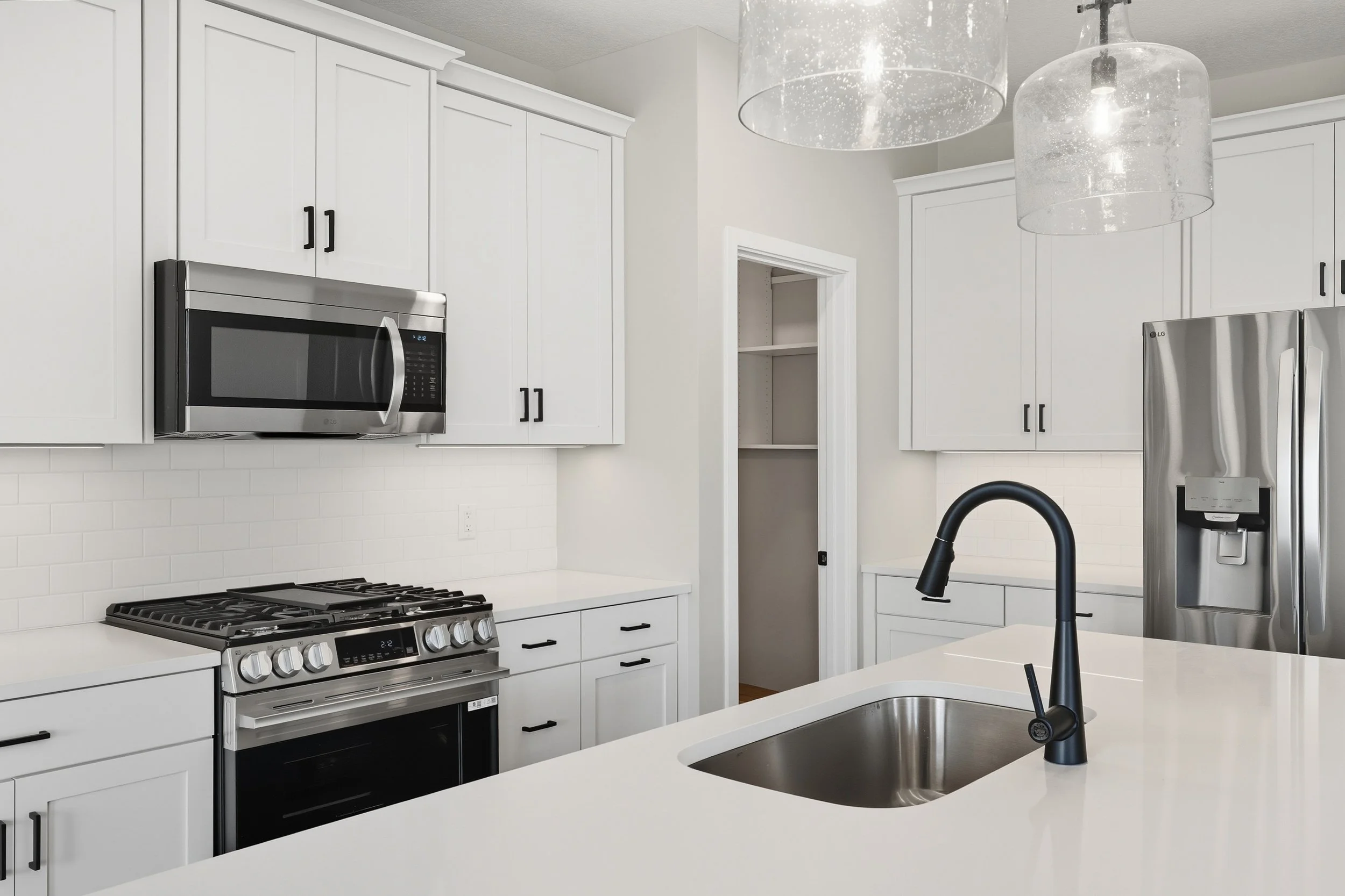 Modern kitchen with white cabinets, stainless steel microwave and refrigerator, black faucet, and white countertops, featuring pendant lights.