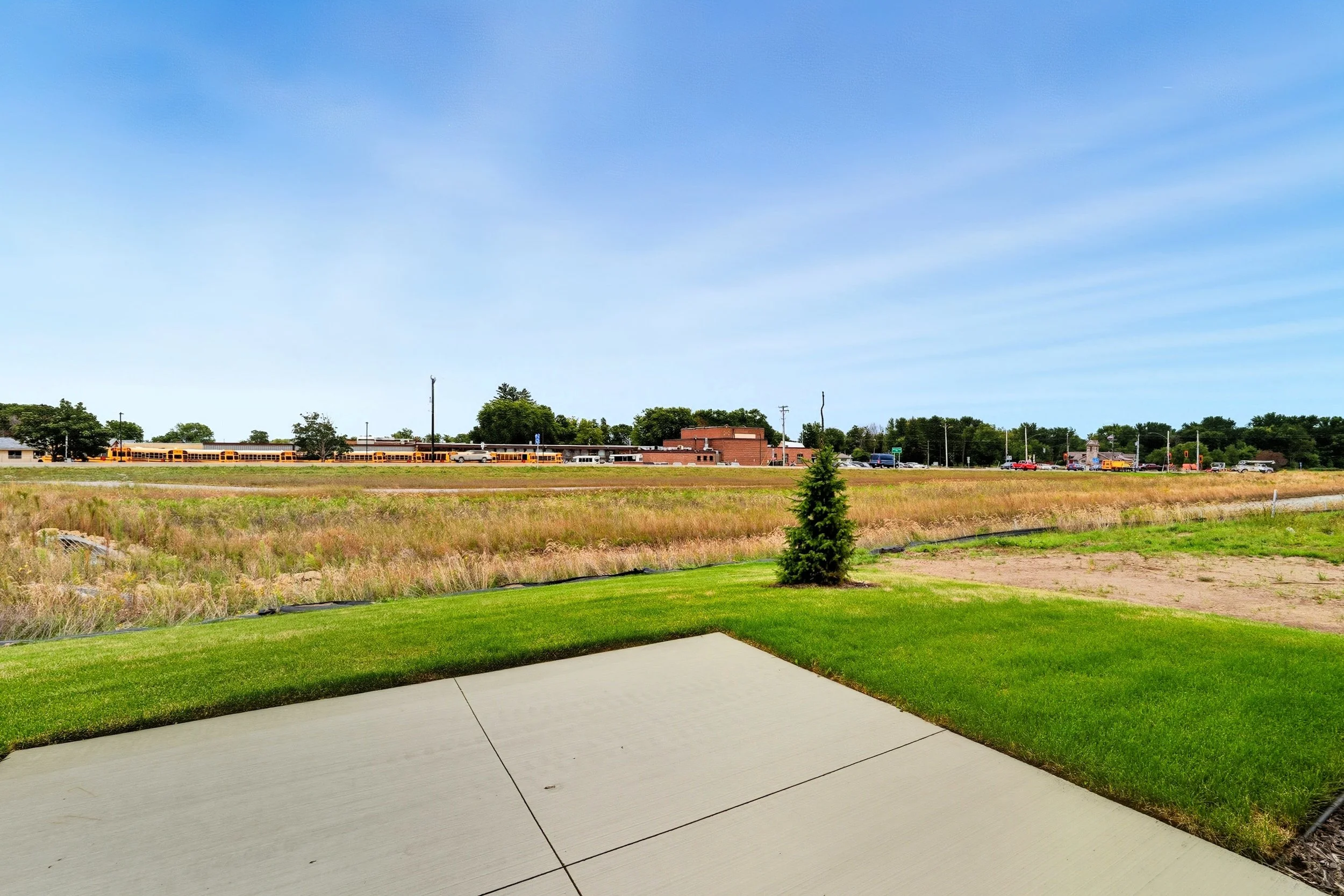 View of a green lawn with a small tree in the foreground, a grassy field, and a building and a yellow school bus in the background under a blue sky.