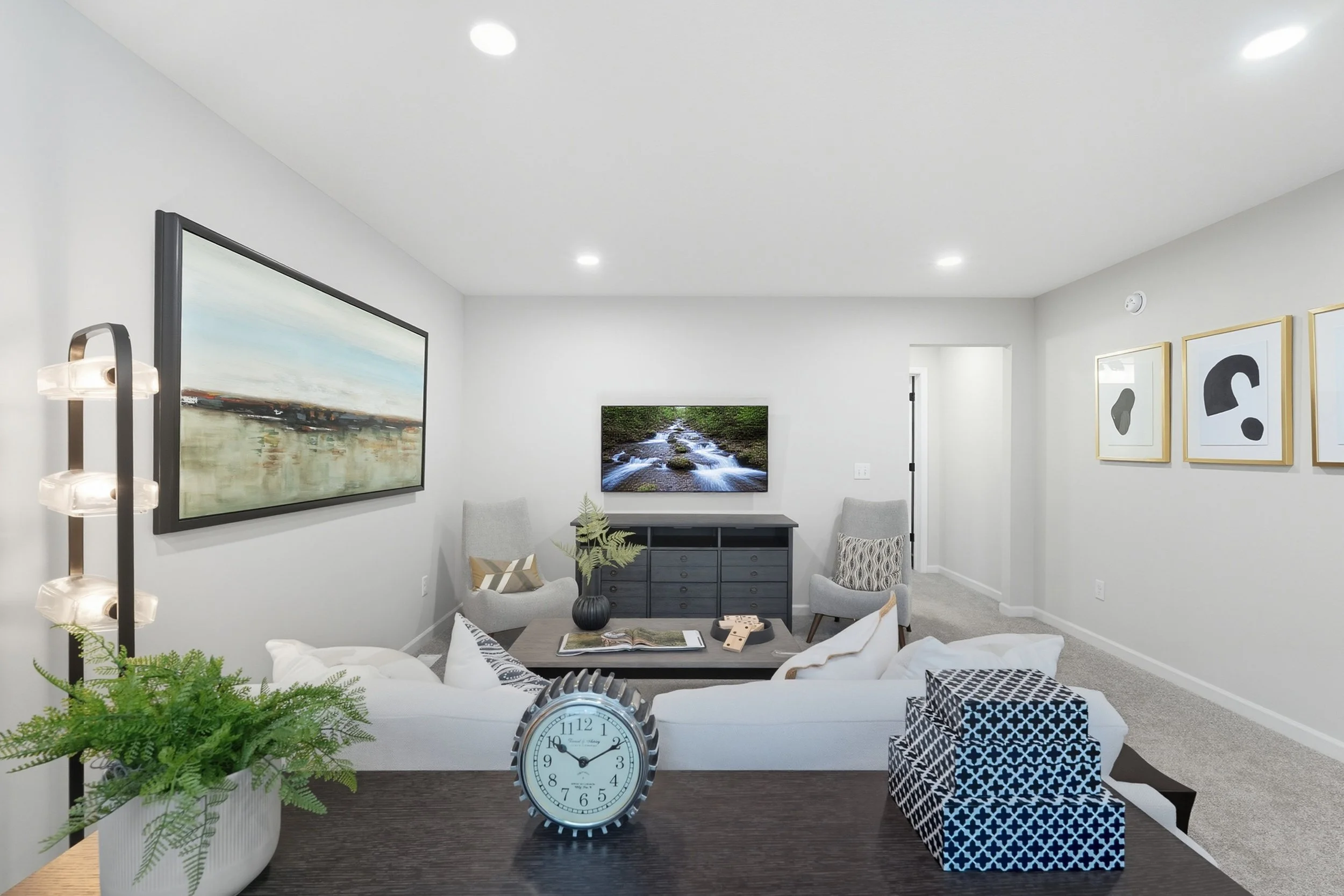 Living room with white sofa, black coffee table, potted fern, wall-mounted TV showing a stream, framed abstract art, and decorative boxes.