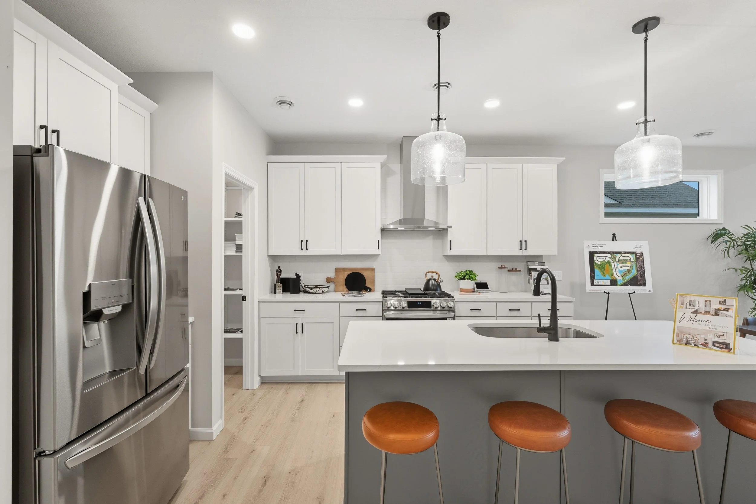 Modern kitchen with white cabinets, stainless steel appliances, light wood flooring, an island with a sink, and brown stools. Pendant lights hang over the island.