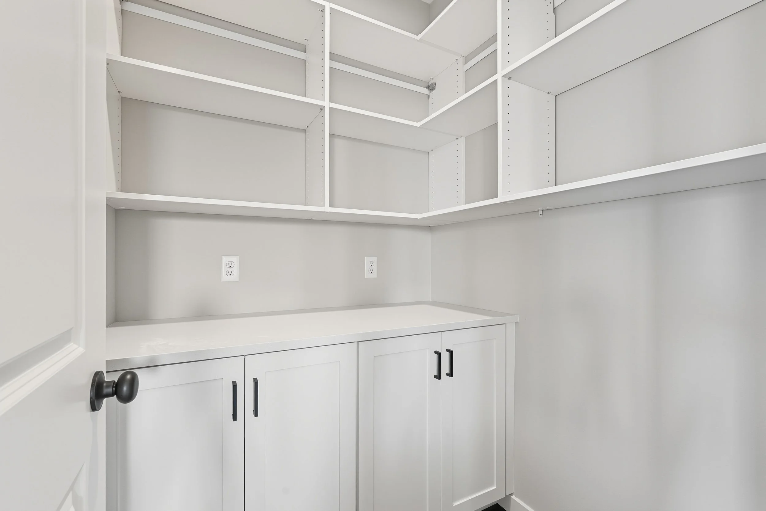 Empty white pantry with open door, white shelving, and cabinets with black handles.