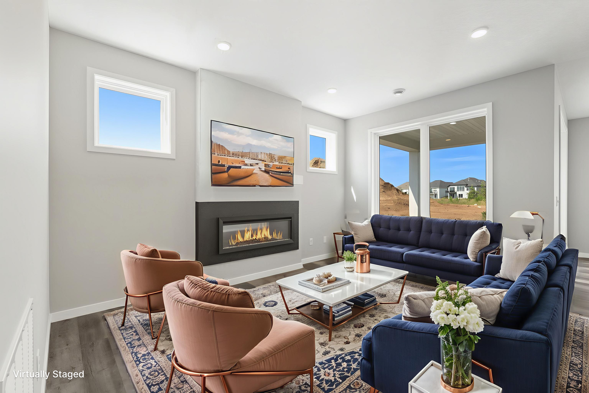 Modern living room with white walls, large windows, a black fireplace, a wall-mounted flat-screen TV, a navy blue sofa, pink accent chairs, a white coffee table, a rug, and views of houses and a hillside outside.