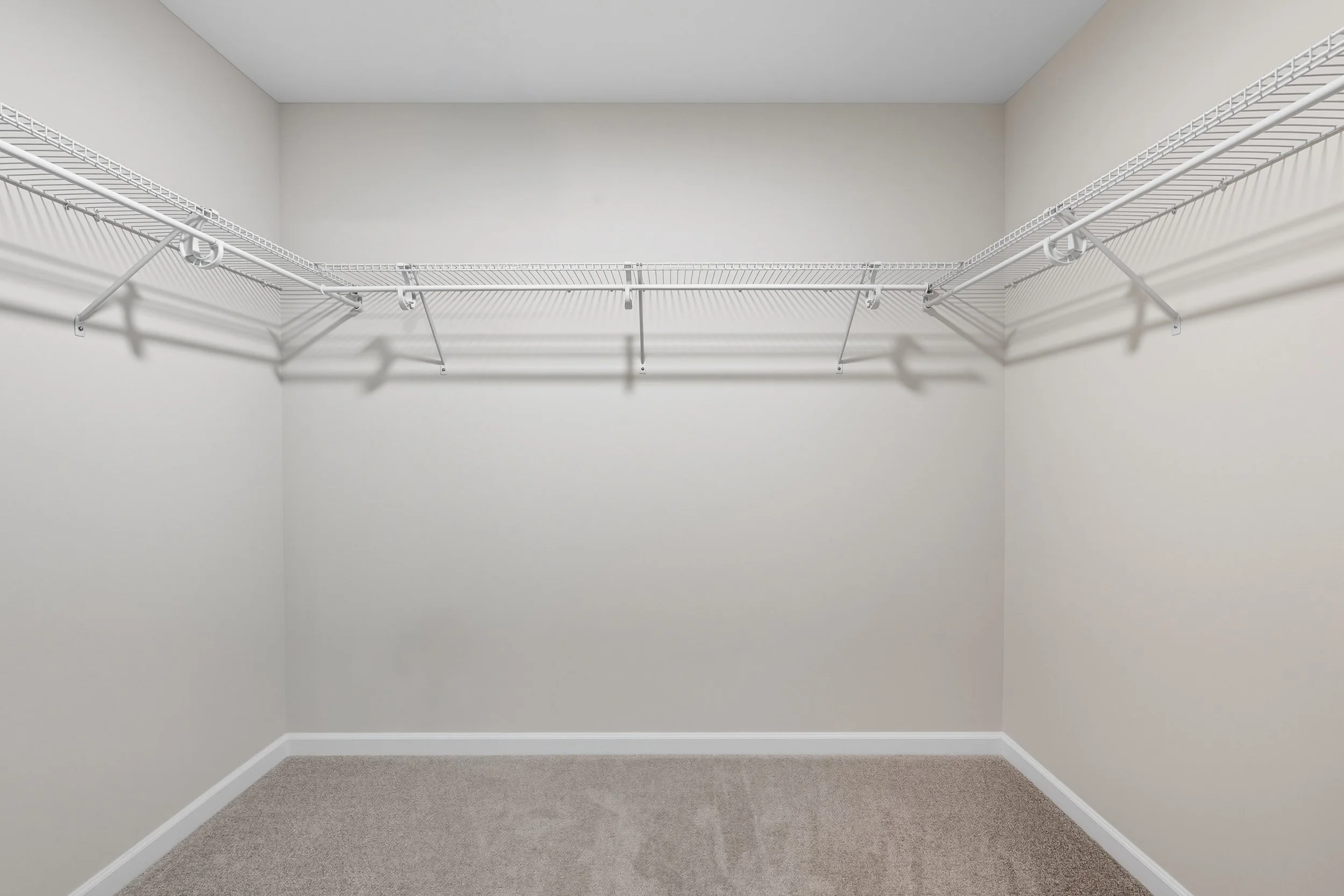 Empty walk-in closet with white wire shelves and closet rod on three walls, beige carpeted floor, and plain light-colored walls.