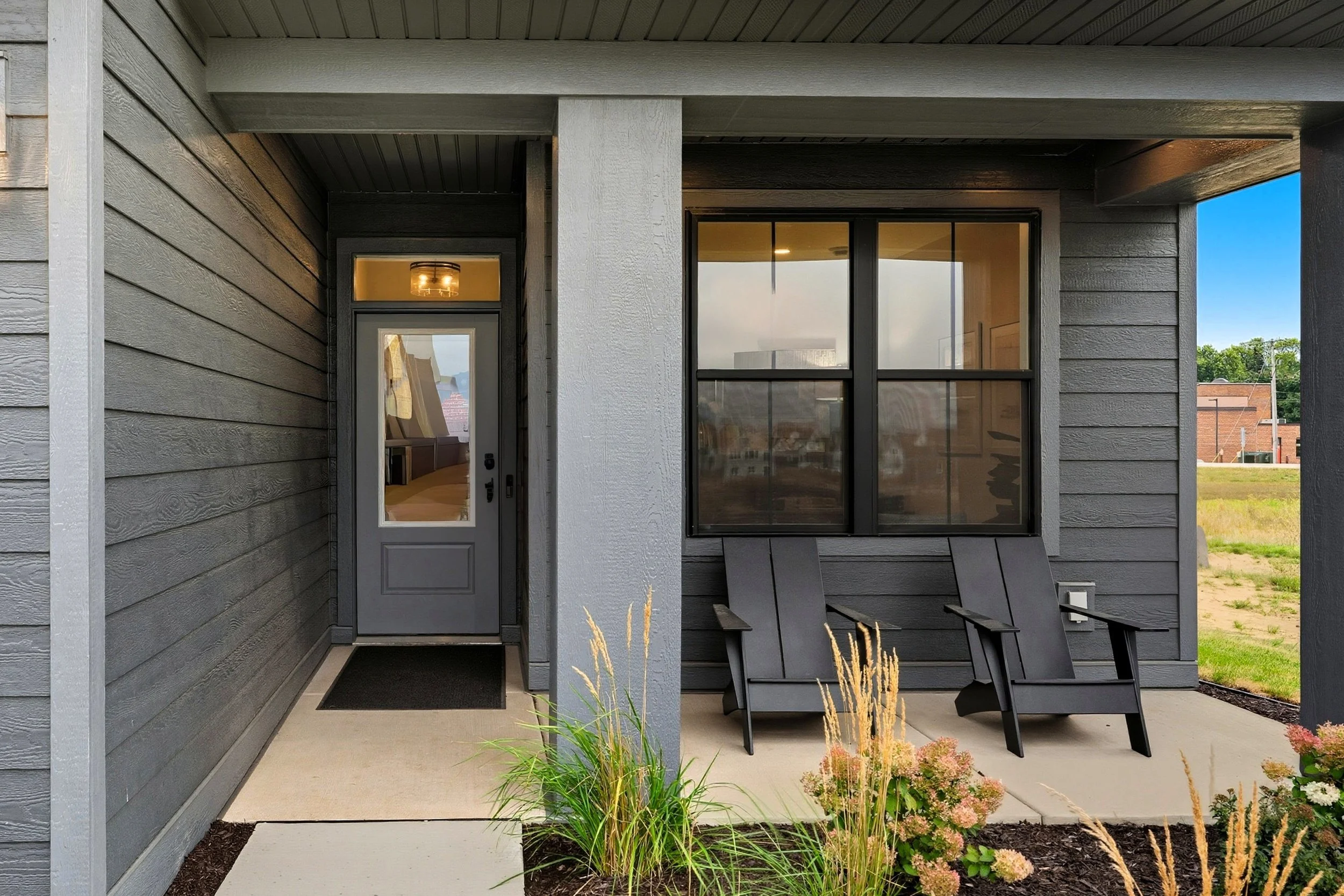 Front porch of a modern house with gray siding, a glass door, two black chairs, and well-maintained landscaping with plants and flowers.