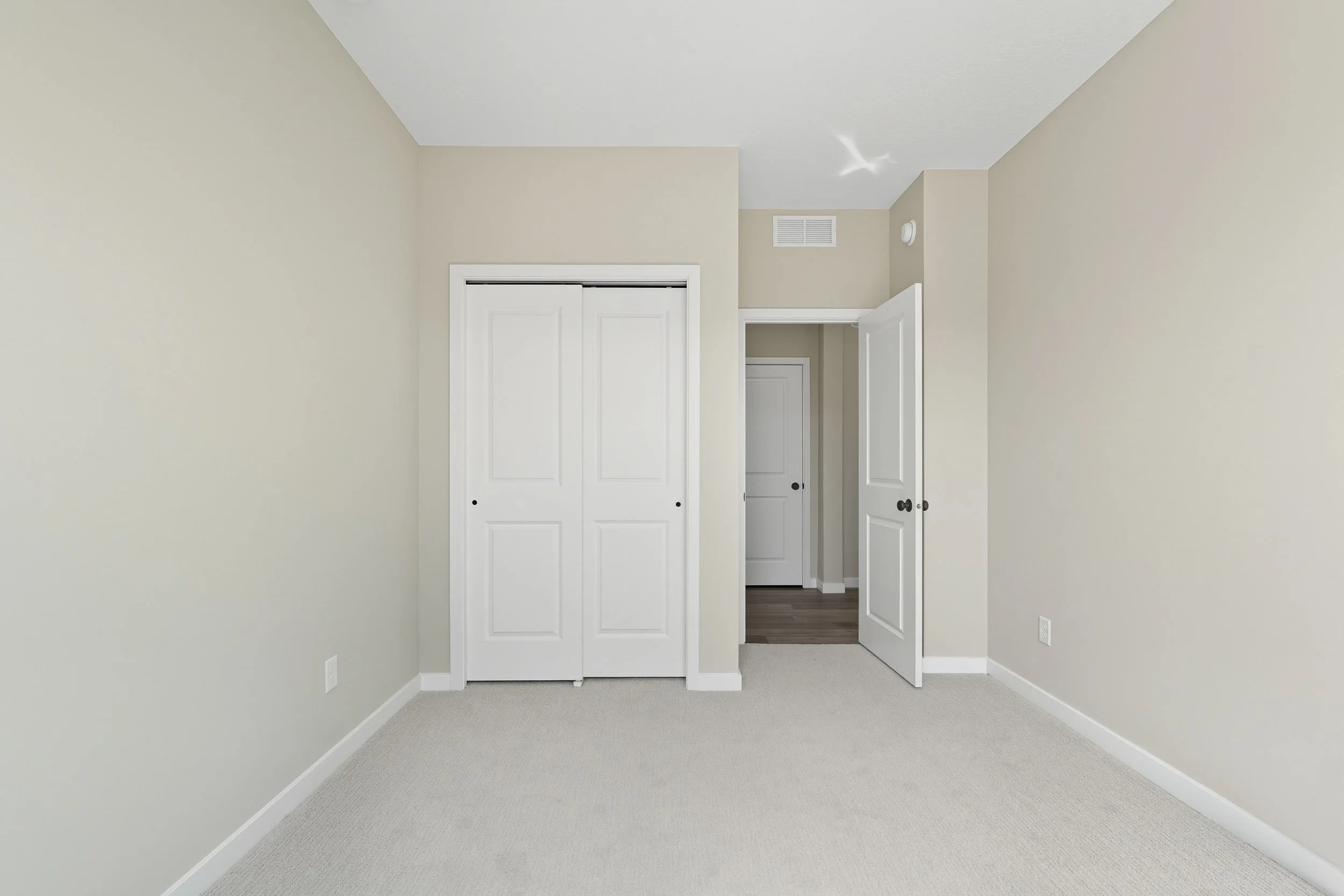 Empty bedroom with beige walls, white doors including closet and doorway, beige carpet, and a view into a hallway with darker wood flooring.