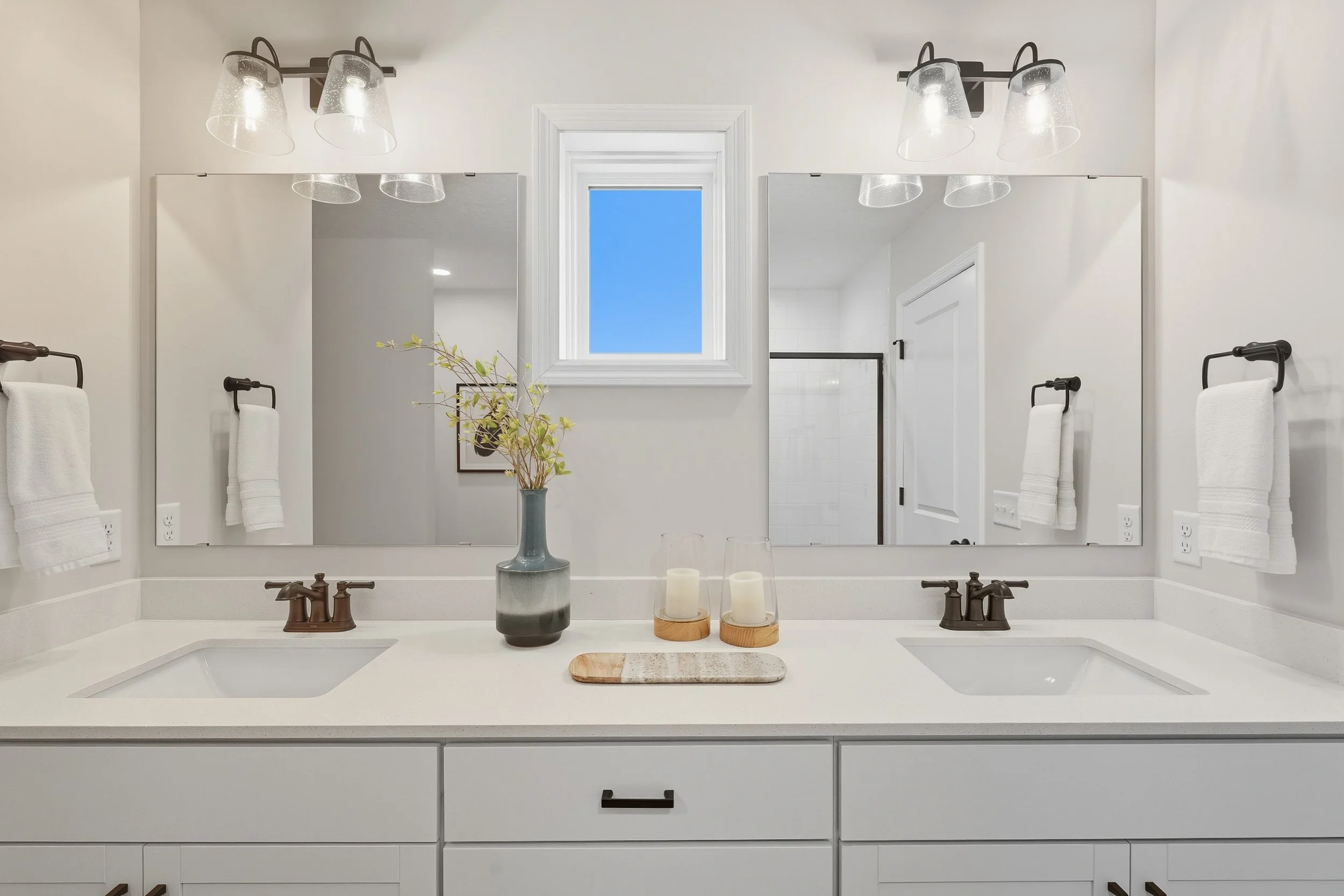 Modern bathroom with white vanity, two sinks, large mirrors, neutral decor, and a small window showing a clear blue sky.