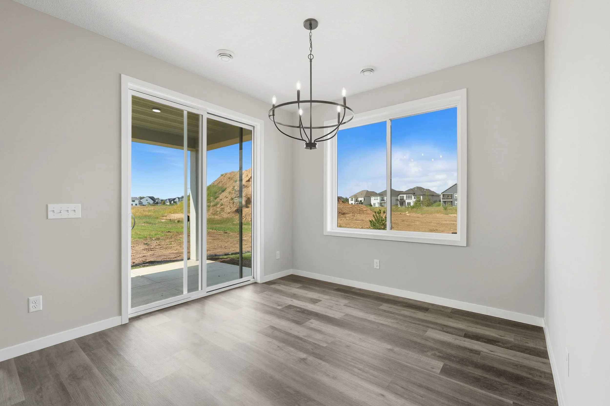 Empty room with wood flooring, a sliding glass door leading to an outdoor patio, and a large window showing houses and a construction site outside, with a modern chandelier hanging from the ceiling.