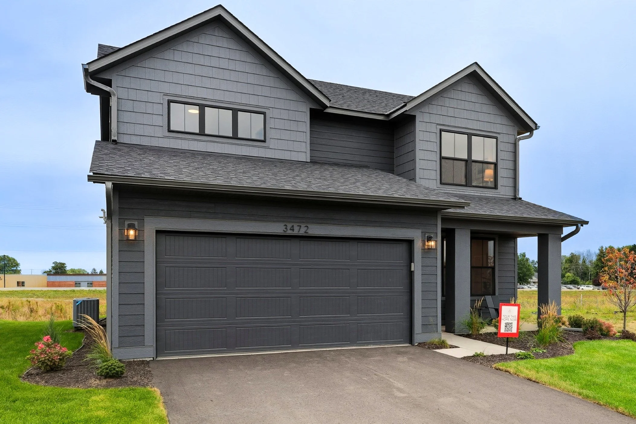 A modern two-story gray house with a dark gray garage door, outdoor lighting, a front porch, and a yard with green grass and landscaped plants.
