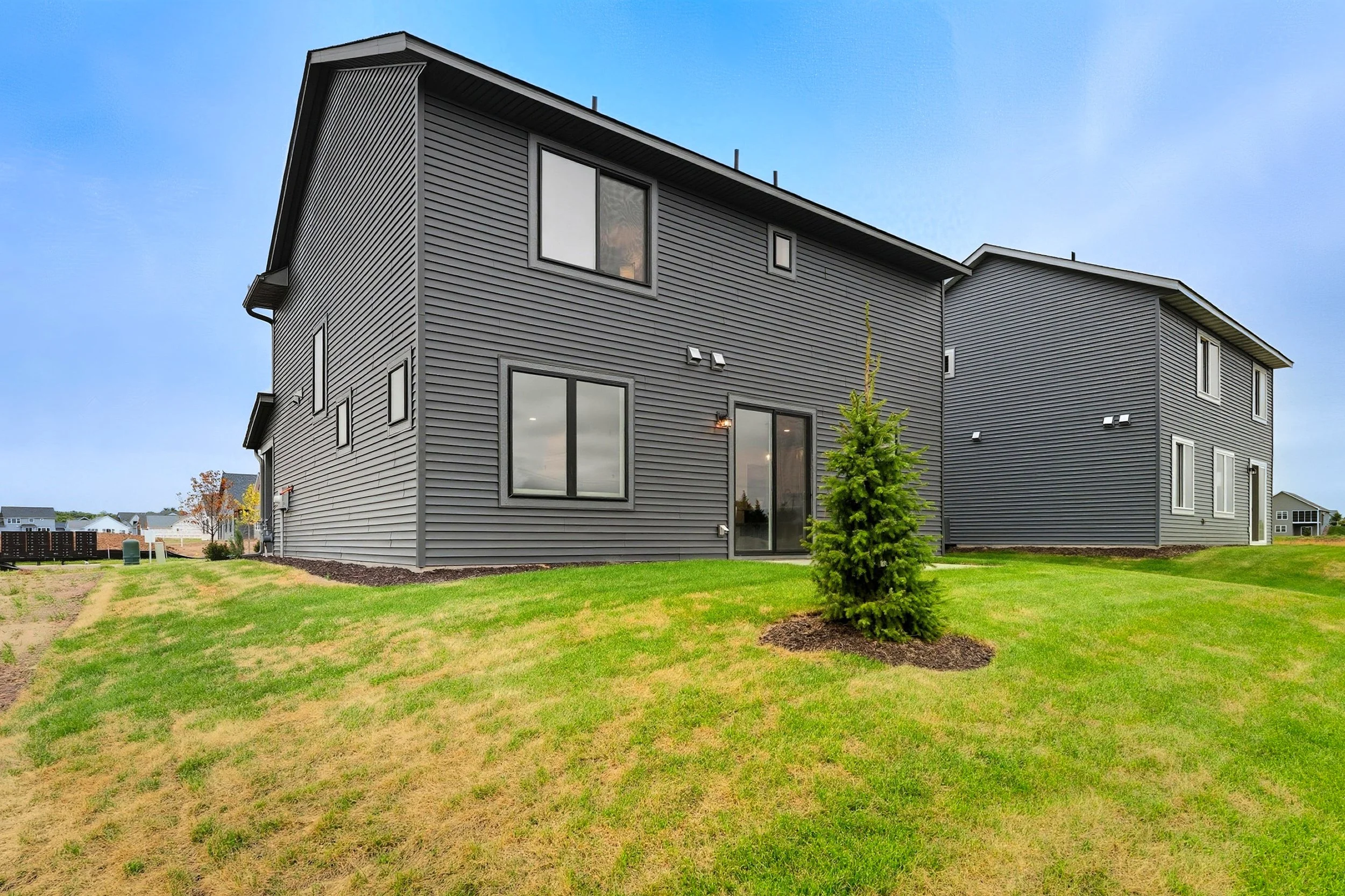 Modern gray two-story house with large windows and a small front lawn with a young pine tree.