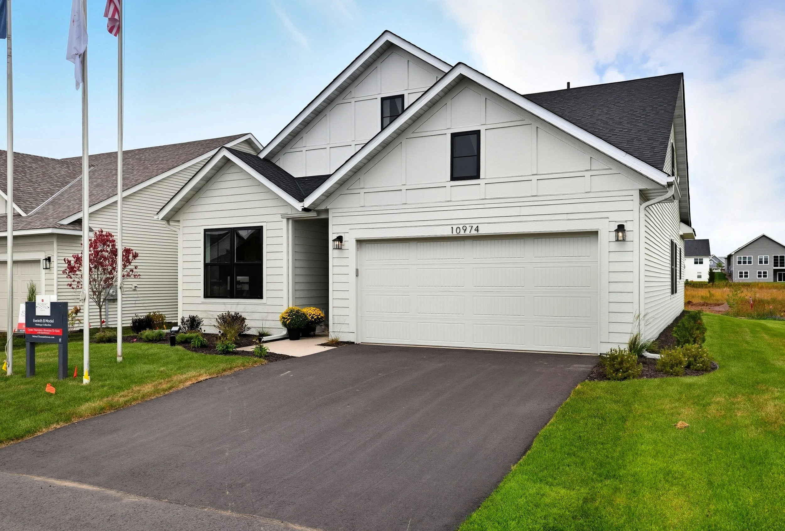 White modern house with black shutters, attached two-car garage, manicured lawn, colorful flower planters, and flags on tall poles in front.