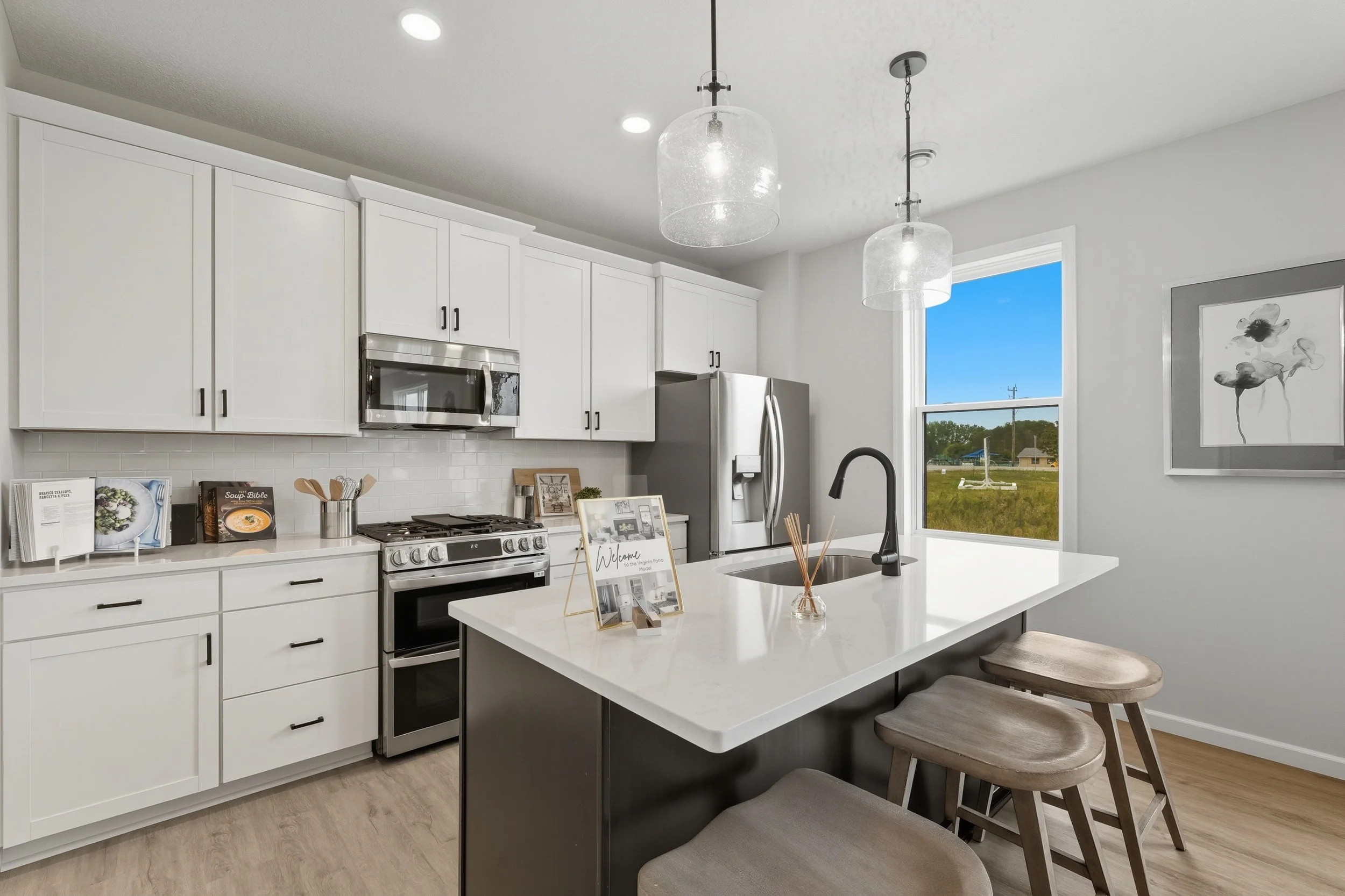 Modern kitchen with white cabinets, stainless steel appliances, a central island with a white countertop, black faucet, and two wooden bar stools. A large window provides natural light, and there are decorative items and cookbooks on the counter.