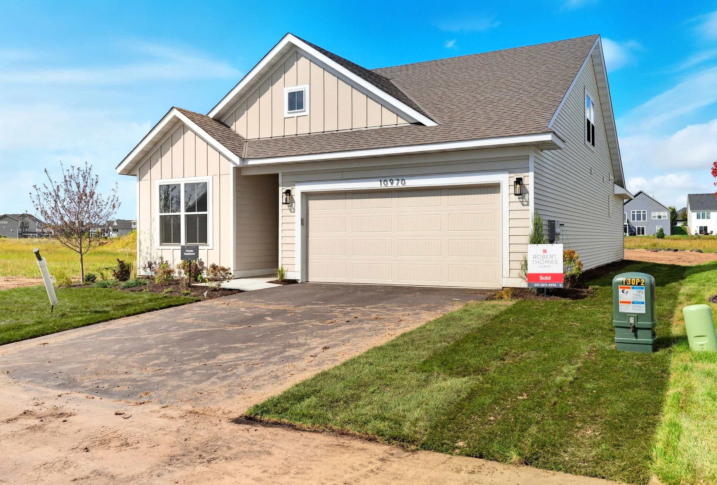 New beige two-story house with a garage, front lawn, and sign indicating it is sold, located in a neighborhood with other houses, under a blue sky.