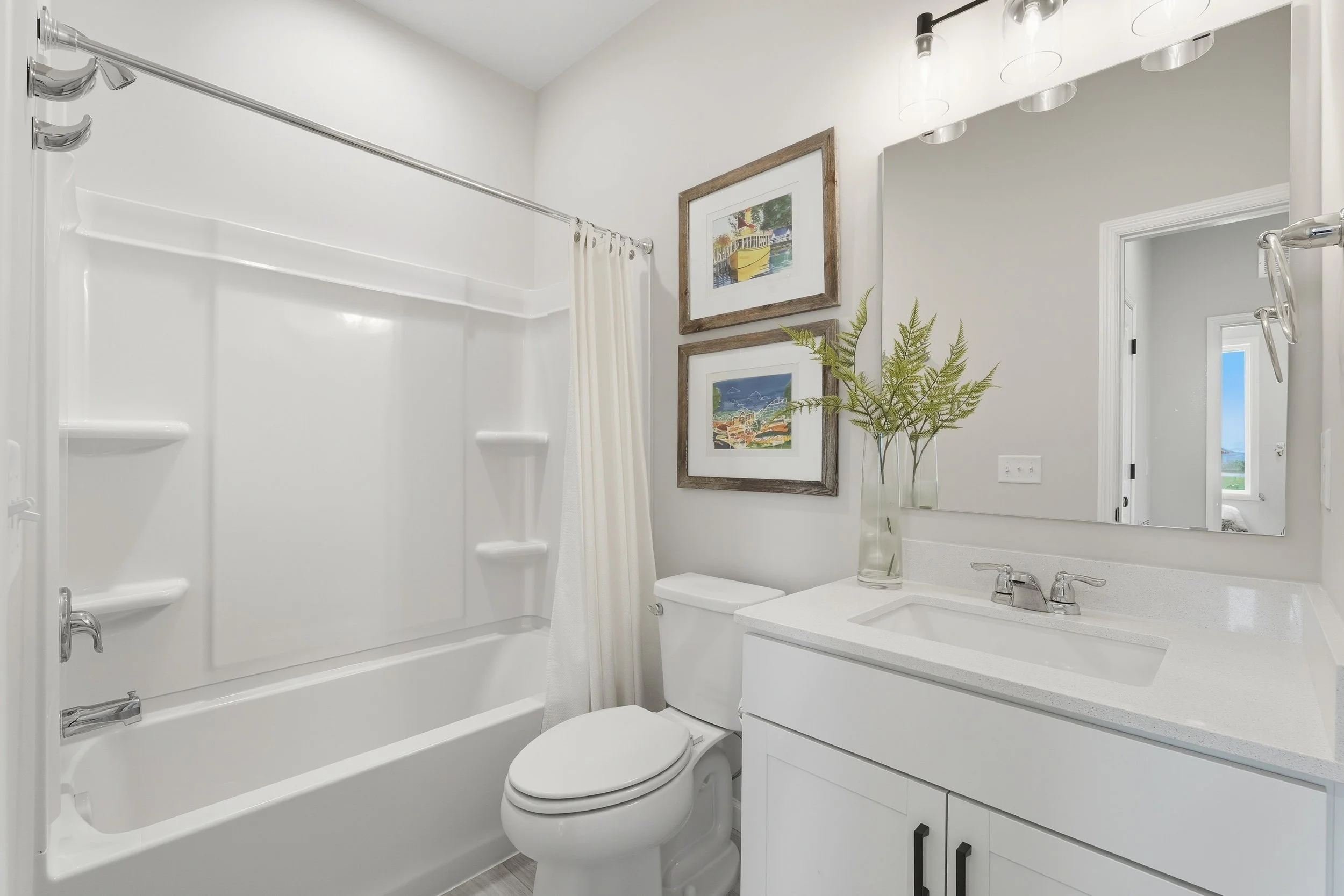 A bathroom with a white vanity, a large mirror, a sink, and a faucet. There are two framed pictures on the wall, a vase with green foliage, a toilet, and a bathtub with a shower curtain rod and curtains.