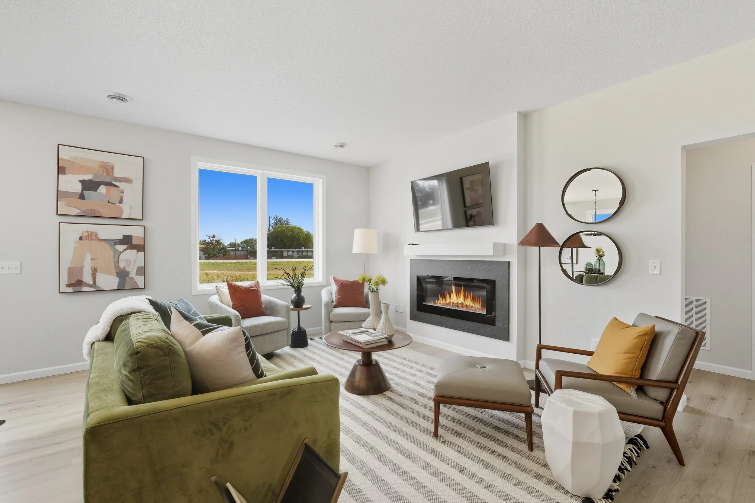 Bright living room with white walls and large window, featuring a green sofa, beige armchairs, a fireplace with a mounted TV above, and decorative mirrors on the wall.
