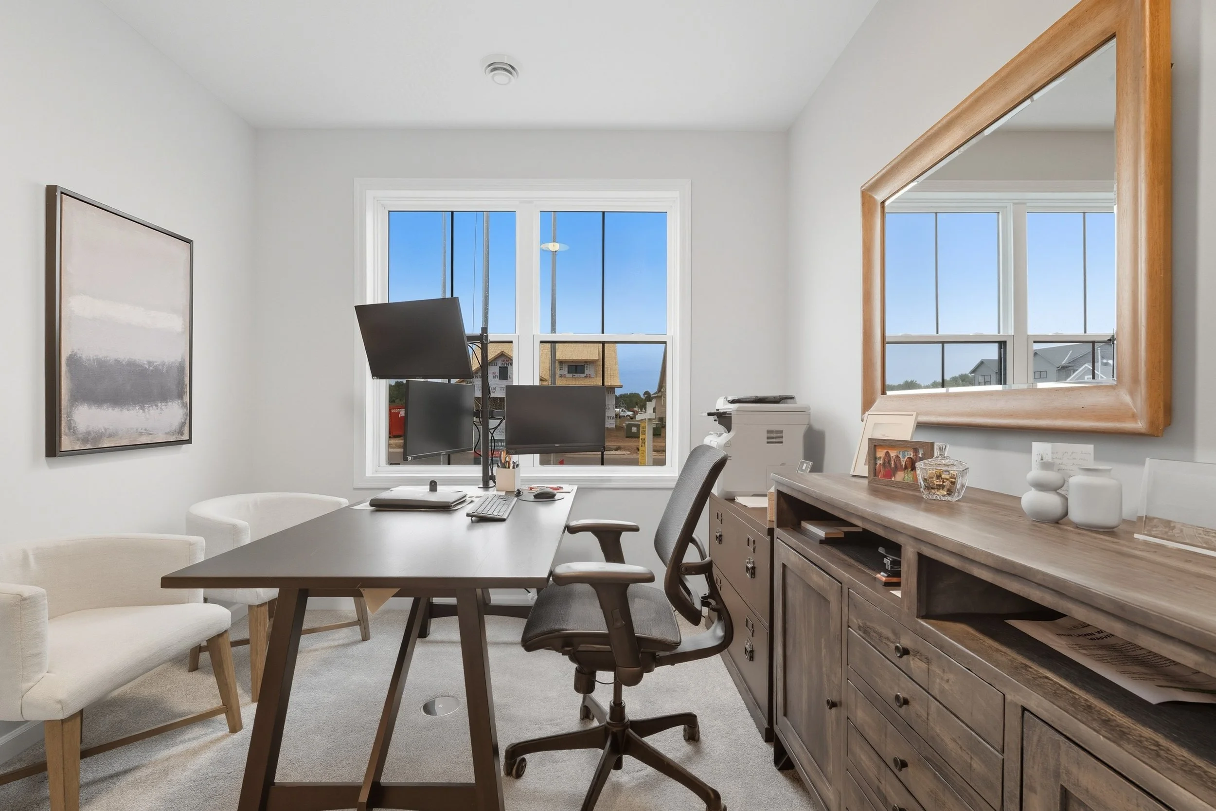 Home office with a wooden desk and black monitors, office chairs, a white armchair, a wooden sideboard with decor, a large mirror, and a window showing neighboring houses and a blue sky.