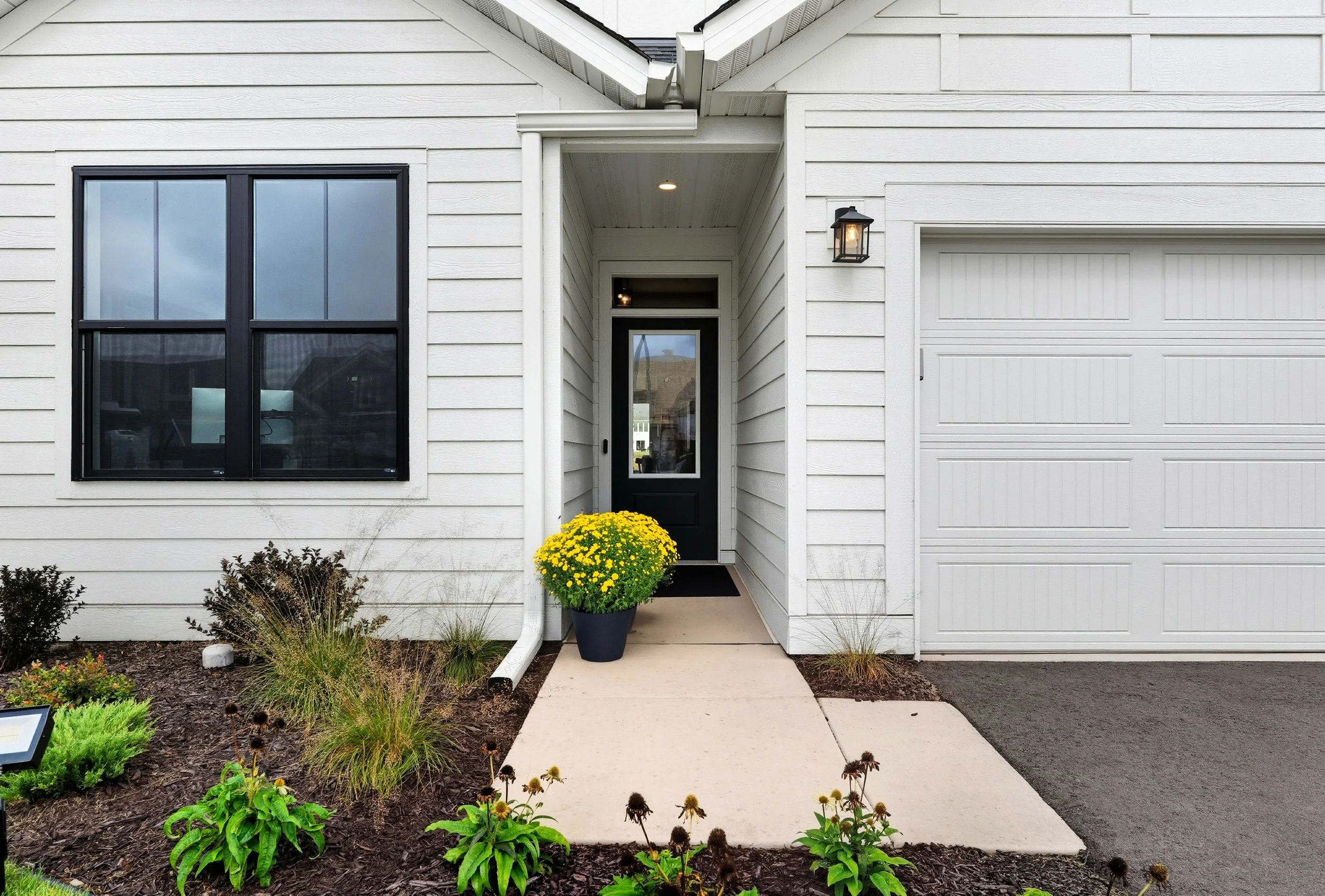 White house entrance with black door, yellow flowers in pot, window to the left, and garage door to the right. Front garden with plants and bushes, concrete walkway leading to the door, outdoor light fixture above the door.
